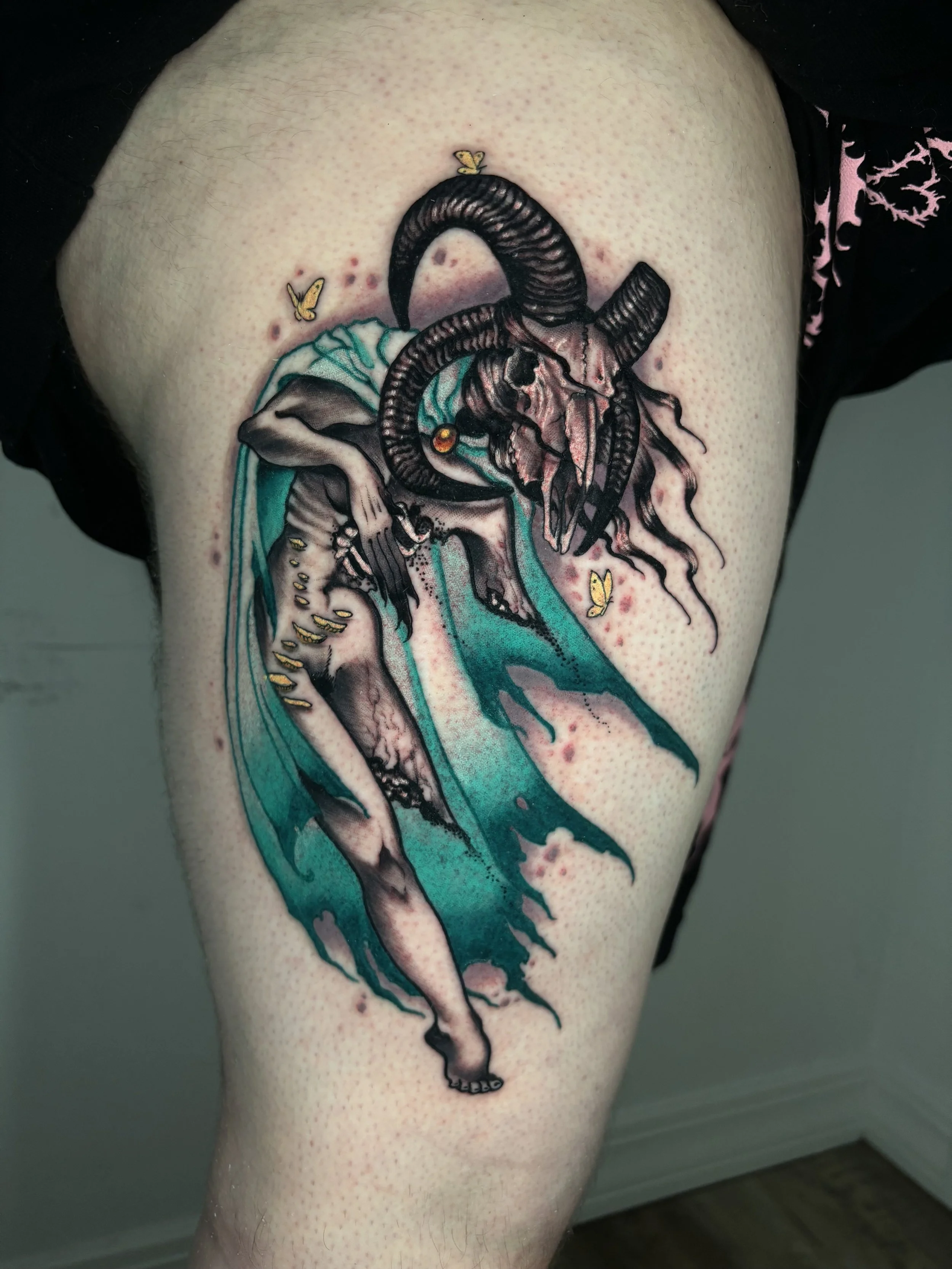 A detailed tattoo of a ram's skull with large curved horns, featuring turquoise and black shading, on a person's upper arm. There are small butterfly stickers around the tattoo.