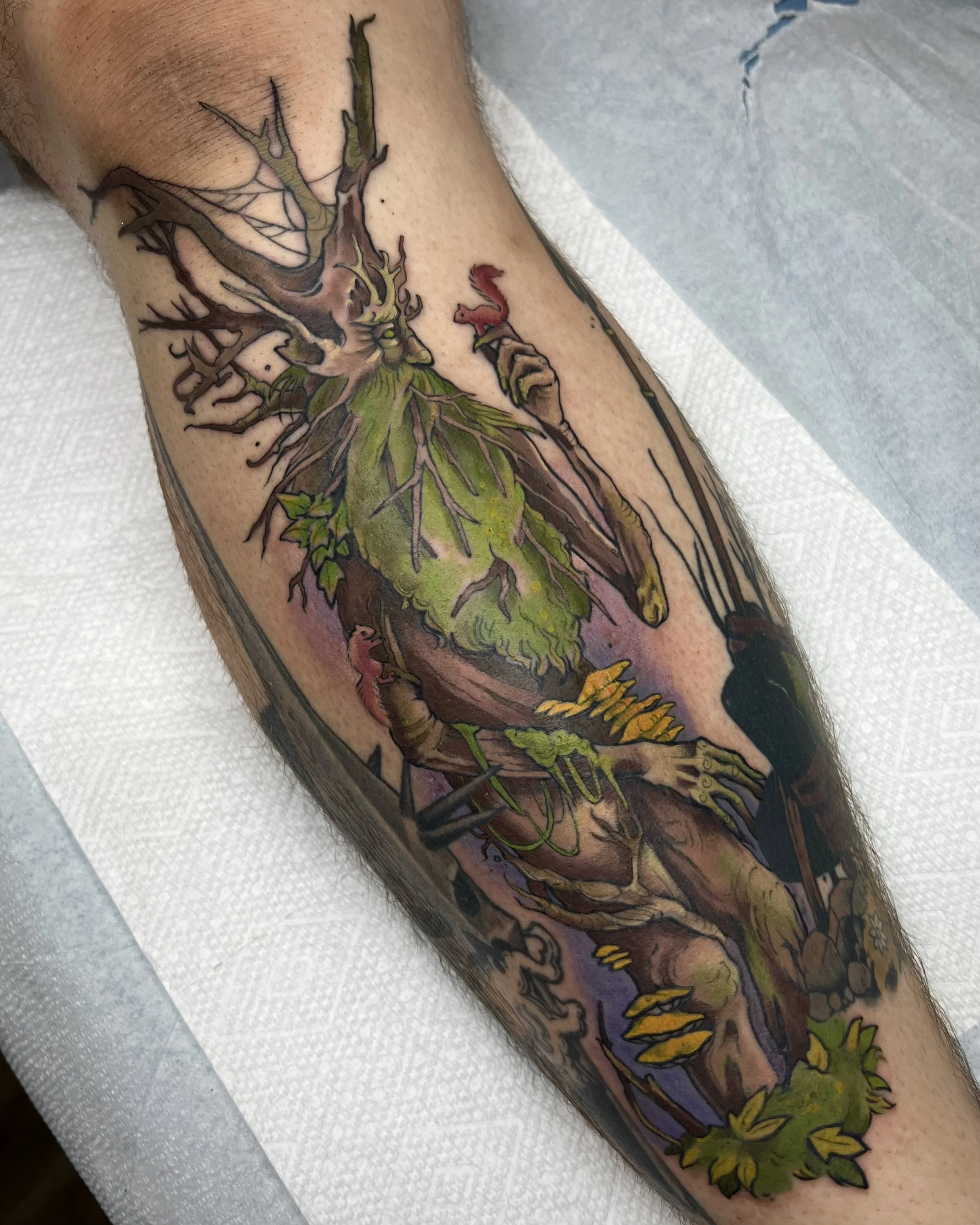 Colorful tattoo on a person's arm featuring a tree with roots, a hand holding a small pink squirrel, and various leaves and flowers. Artwork includes green, brown, yellow, and purple hues.