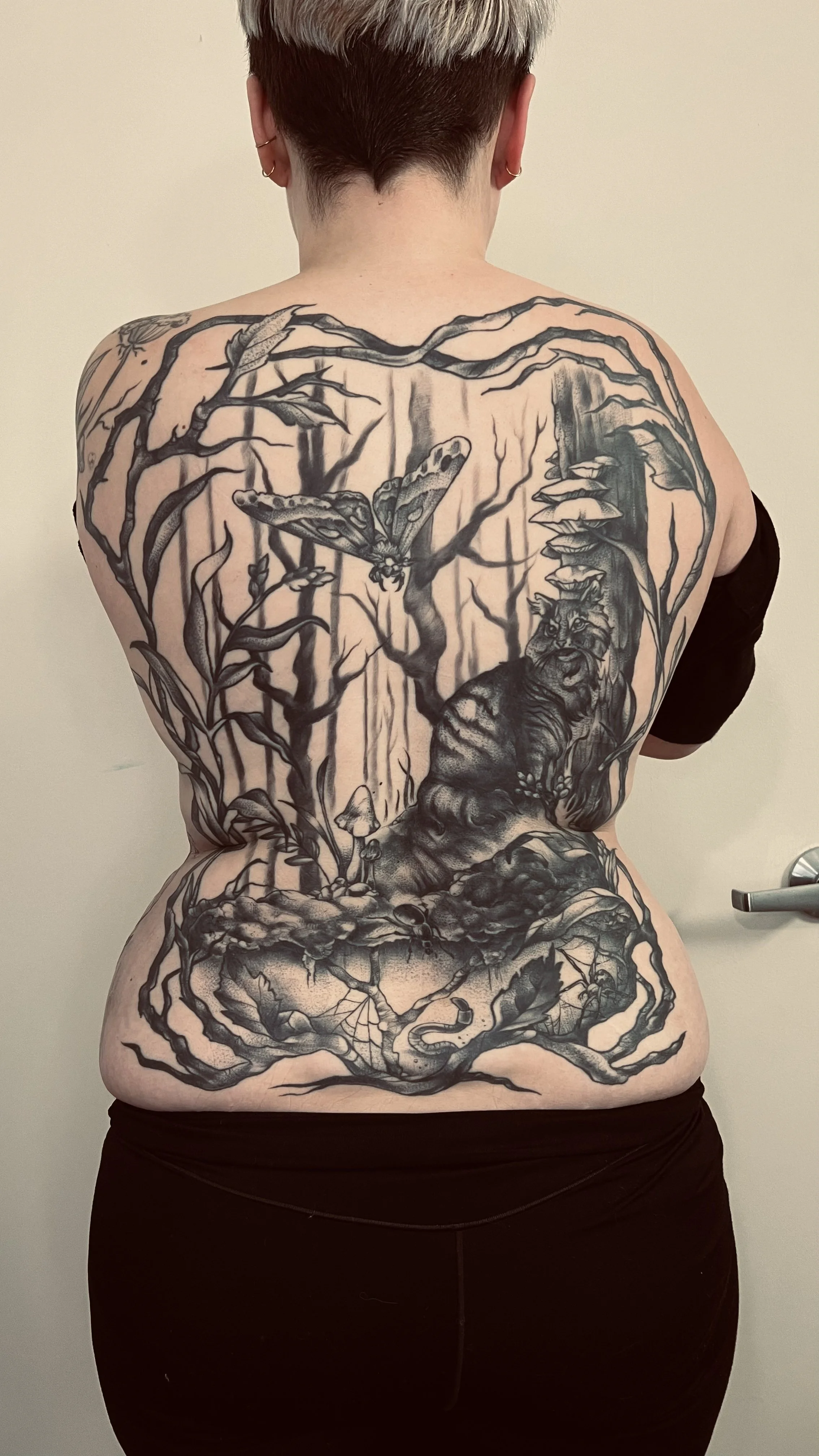 Back view of a person with short, multicolored hair and earrings, showcasing a large black and gray nature-themed tattoo that covers the entire back, featuring a lion, trees, a moth, mushrooms, and various plants.