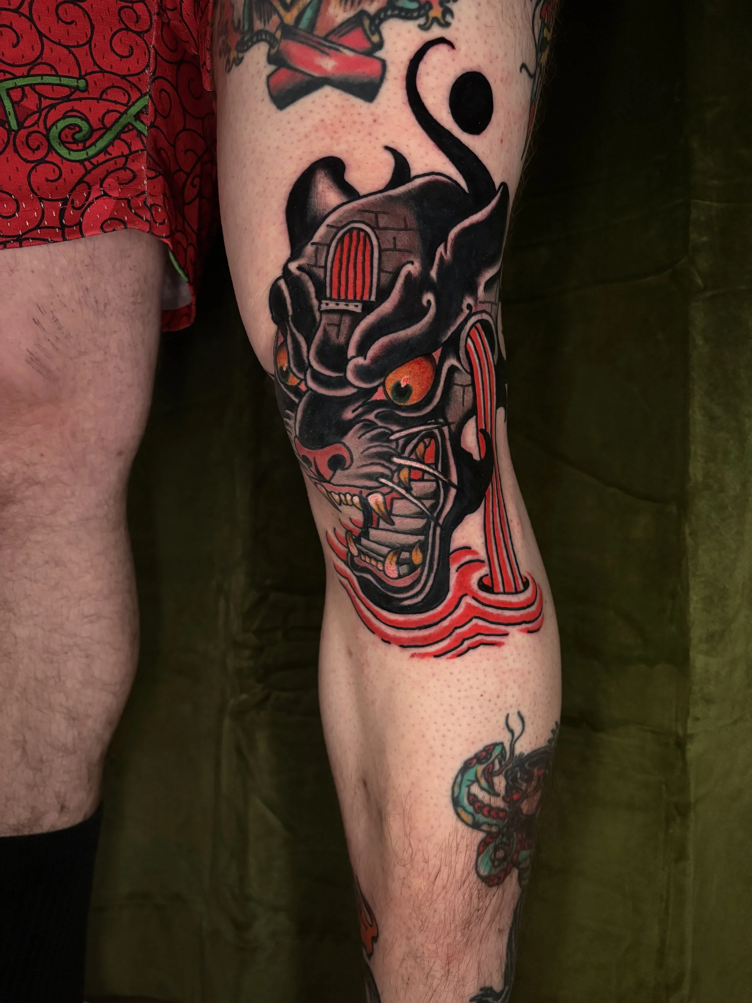 A tattoo on a person's calf features a detailed black and red dragon with glowing orange eyes, surrounded by red and black elements, including a skull-like face and flowing lines.