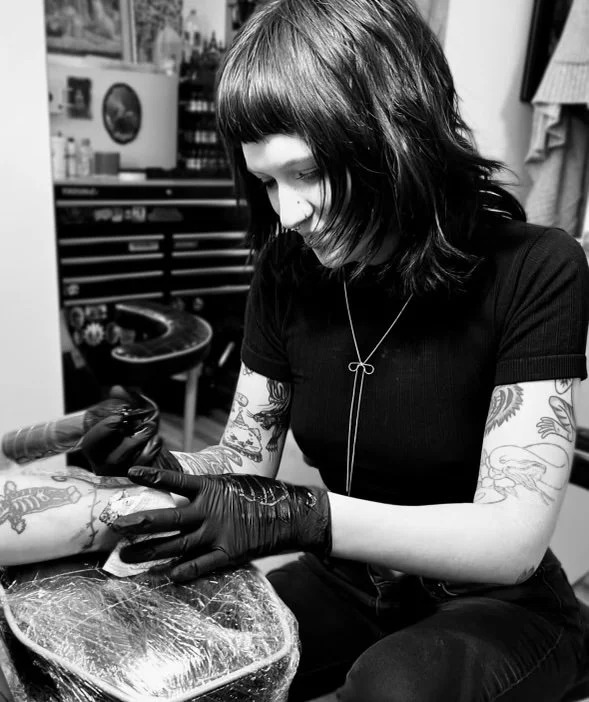 Tattoo artist working on a person's arm in a tattoo studio, with tattoo supplies and equipment in the background.