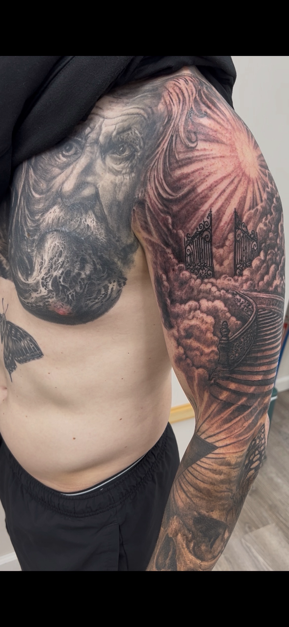 Tattoo of a lion's face on the shoulder with a landscape scene including a gate, clouds, and a staircase on the arm.