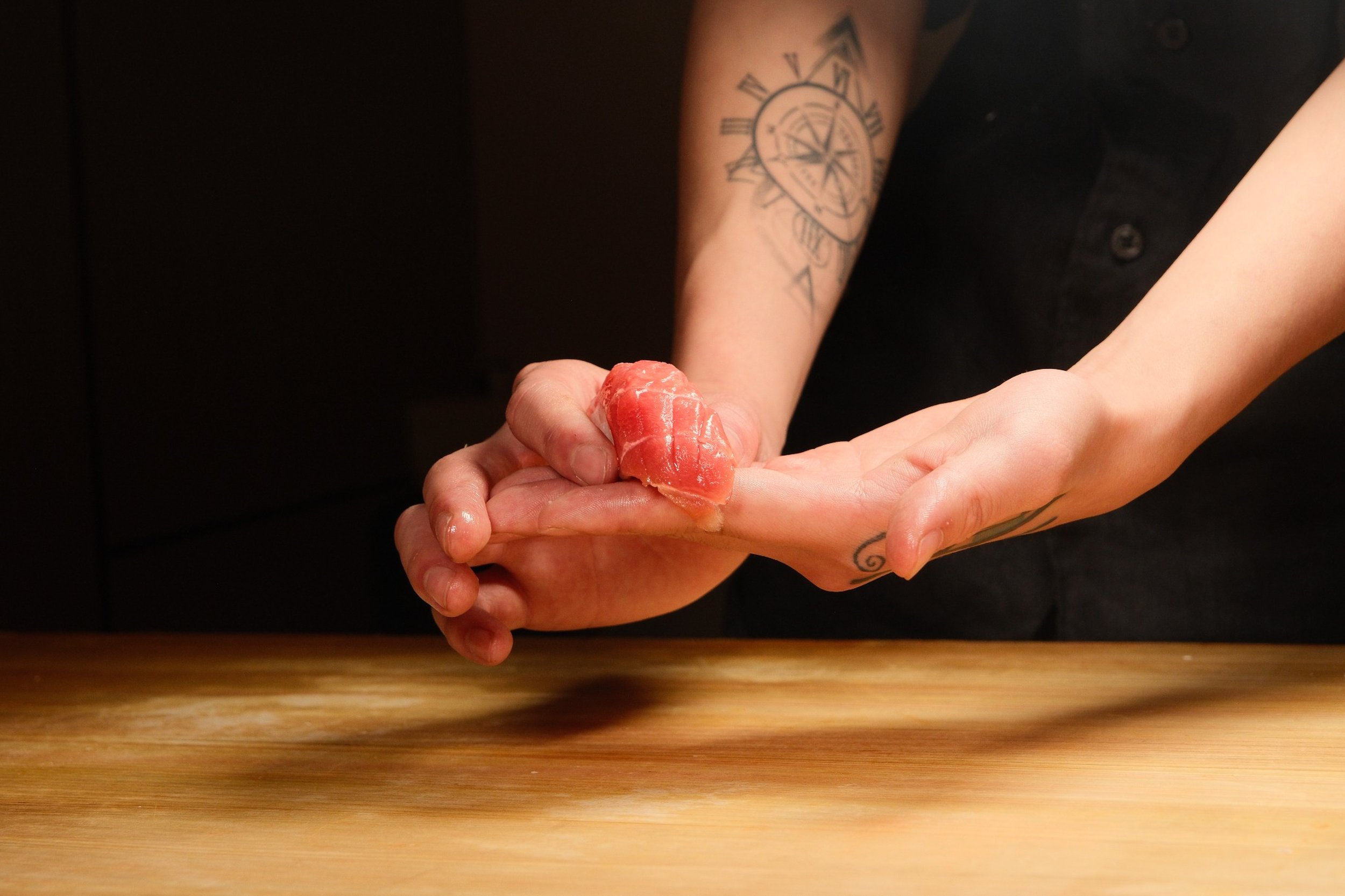 A person with tattoos on their arm holding a piece of raw meat or fish over a wooden surface.