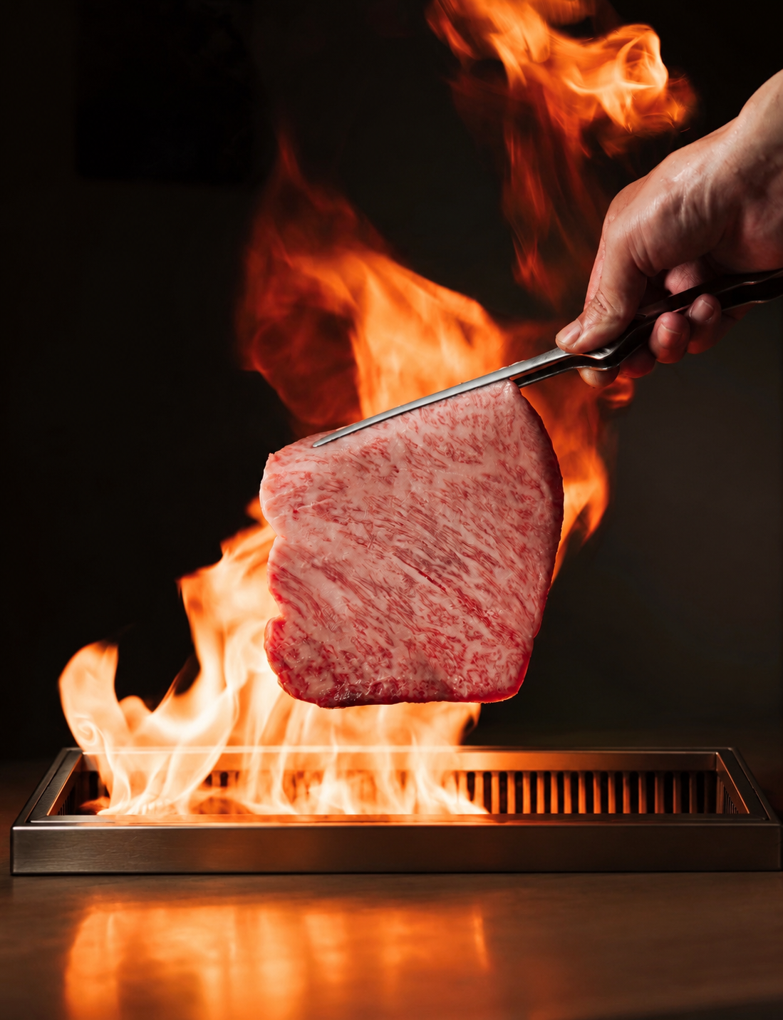 A hand holding tongs with a marbled piece of wagyu beef over an open flame in a grill.