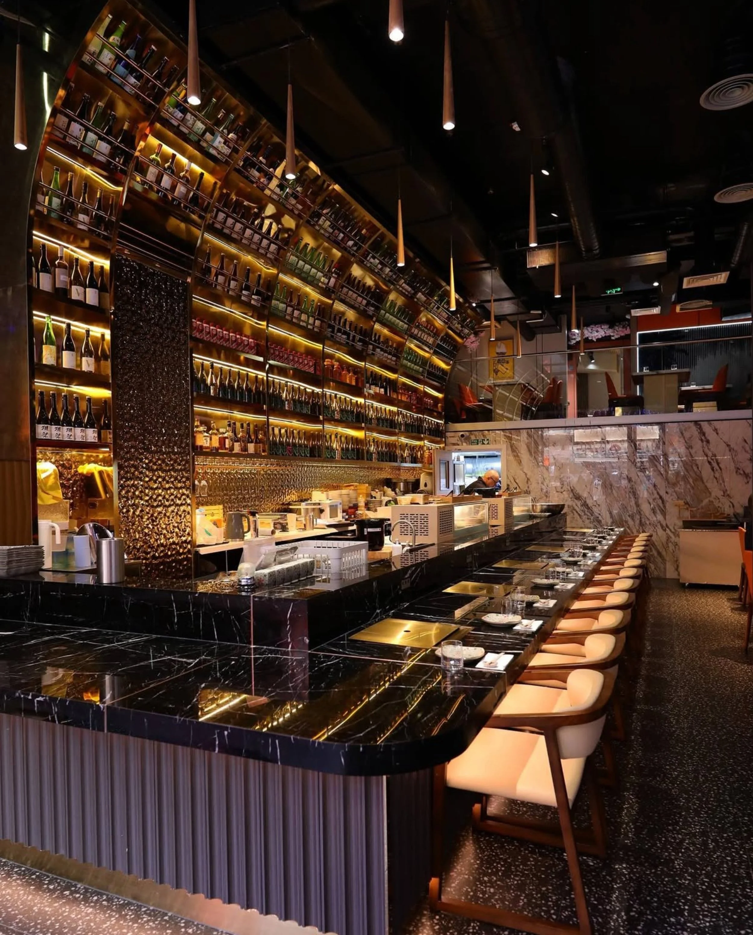 Interior of a modern restaurant with a black marble bar counter, beige chairs, a large illuminated wooden wall with shelves of bottles, and a dark ceiling with hanging pendant lights.