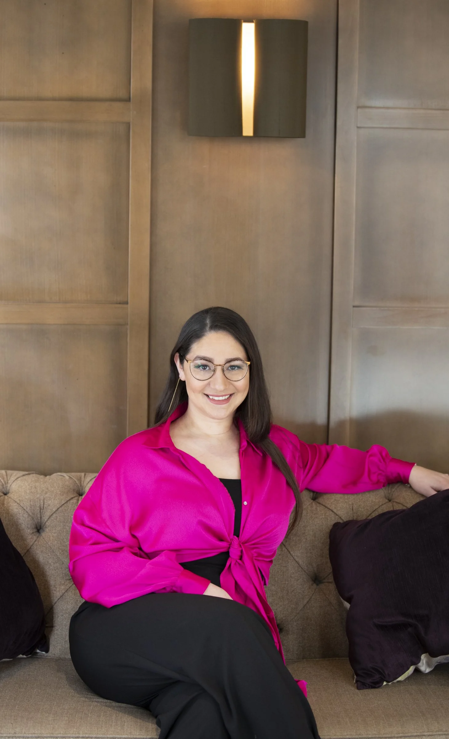 A woman with glasses and long brown hair sitting on a brown couch, wearing a bright pink blouse and black pants, smiling, with a wooden wall and a lit wall sconce behind her.