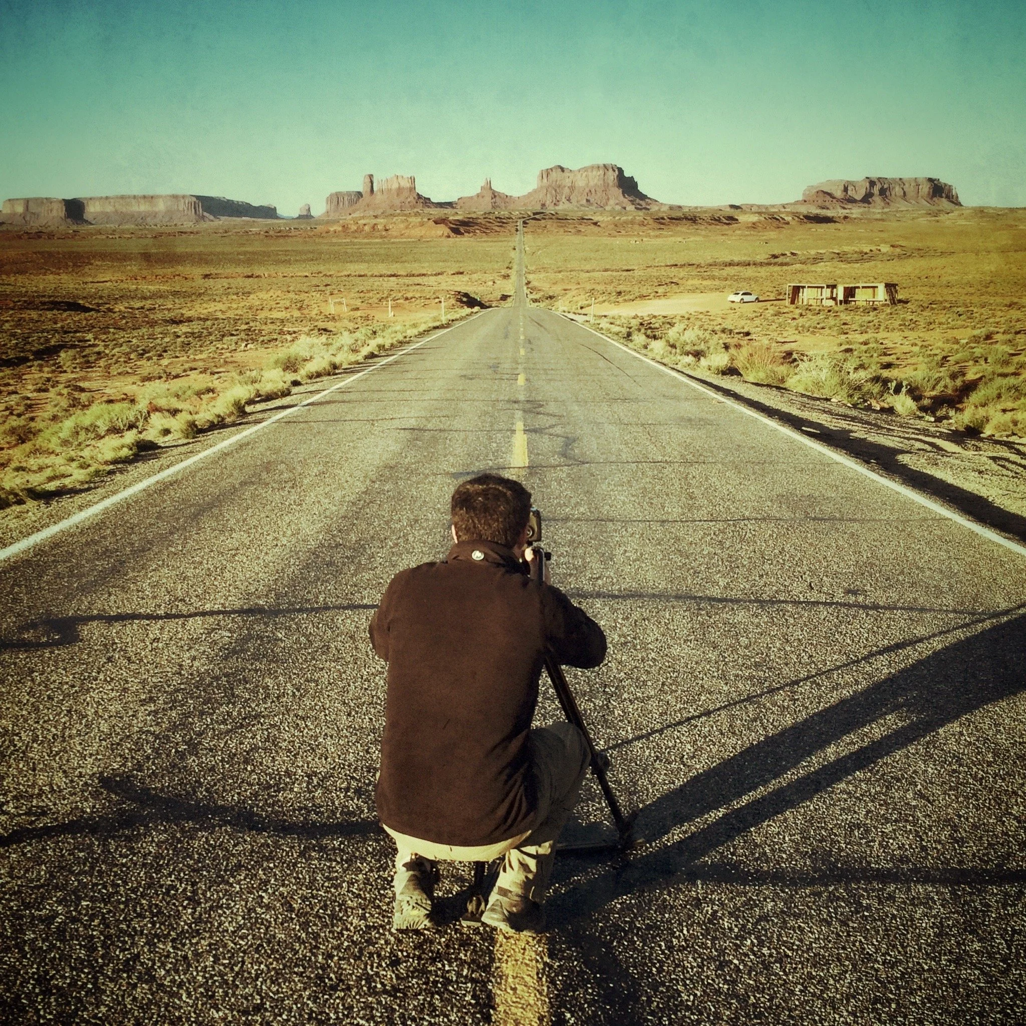 A filmmaker composing a shot along a desert road toward distant sandstone formations
