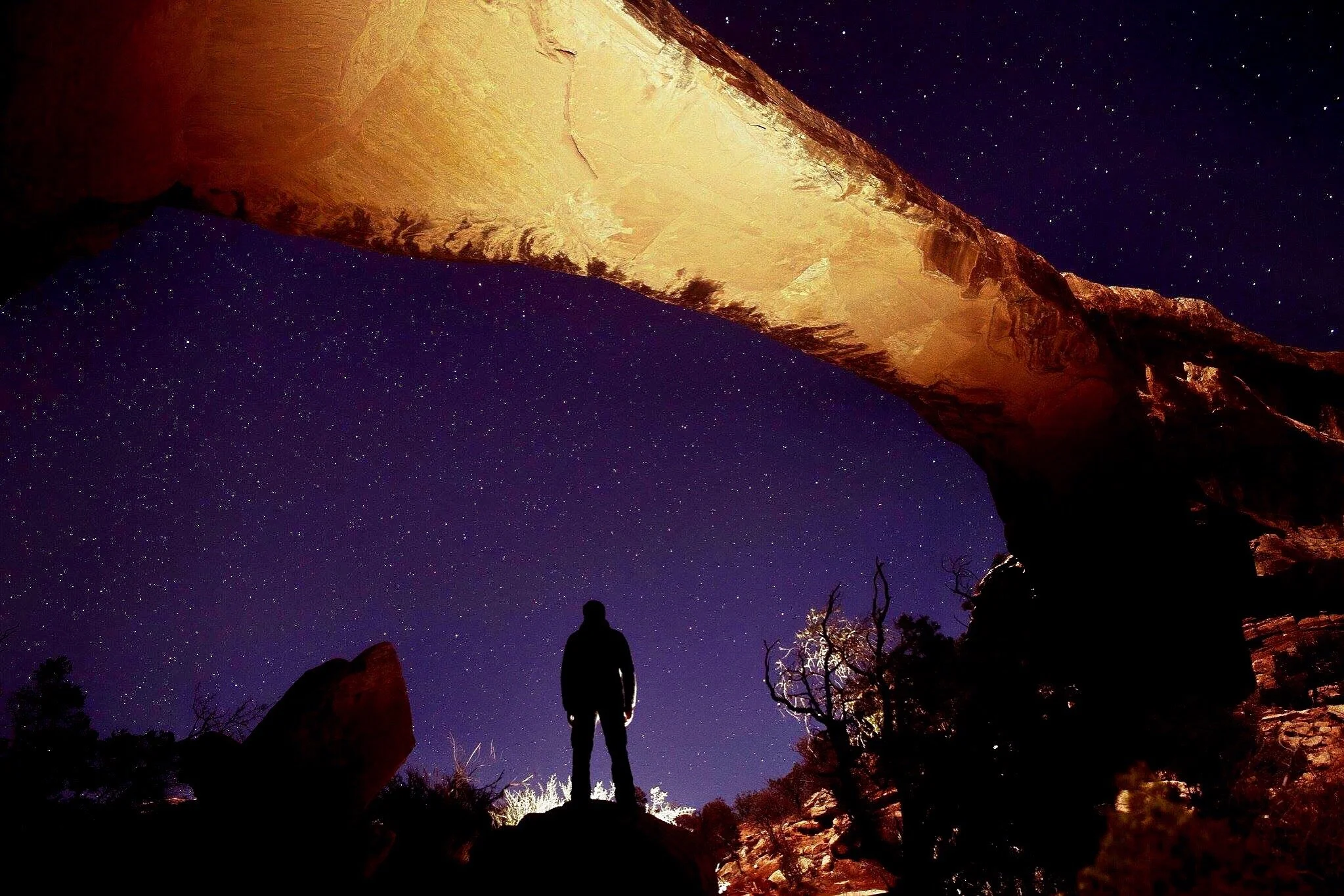 A lone silhouette beneath a sandstone arch and a star-filled sky in Utah.