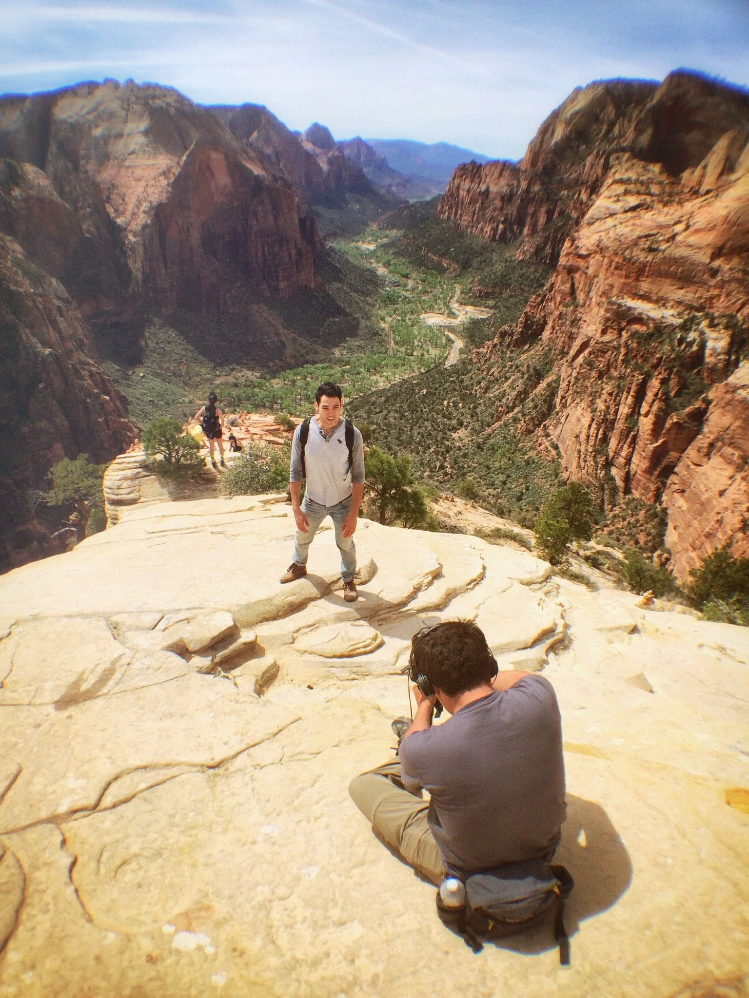 A photographer frames a person standing above a steep canyon, showing the human scale of the landscape.