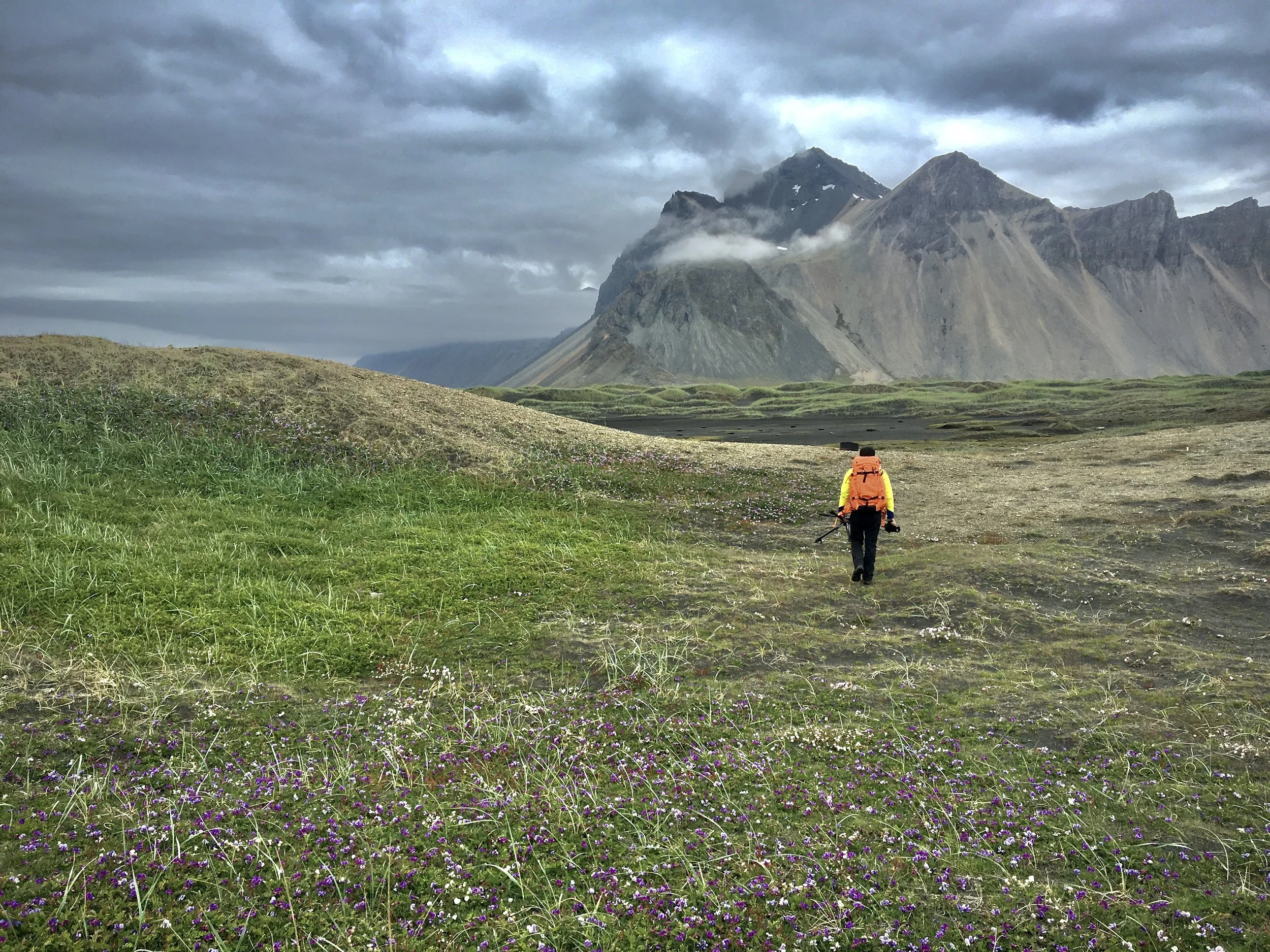 A hiker in an orange jacket crosses green terrain beneath dark clouds and mist-covered mountains.