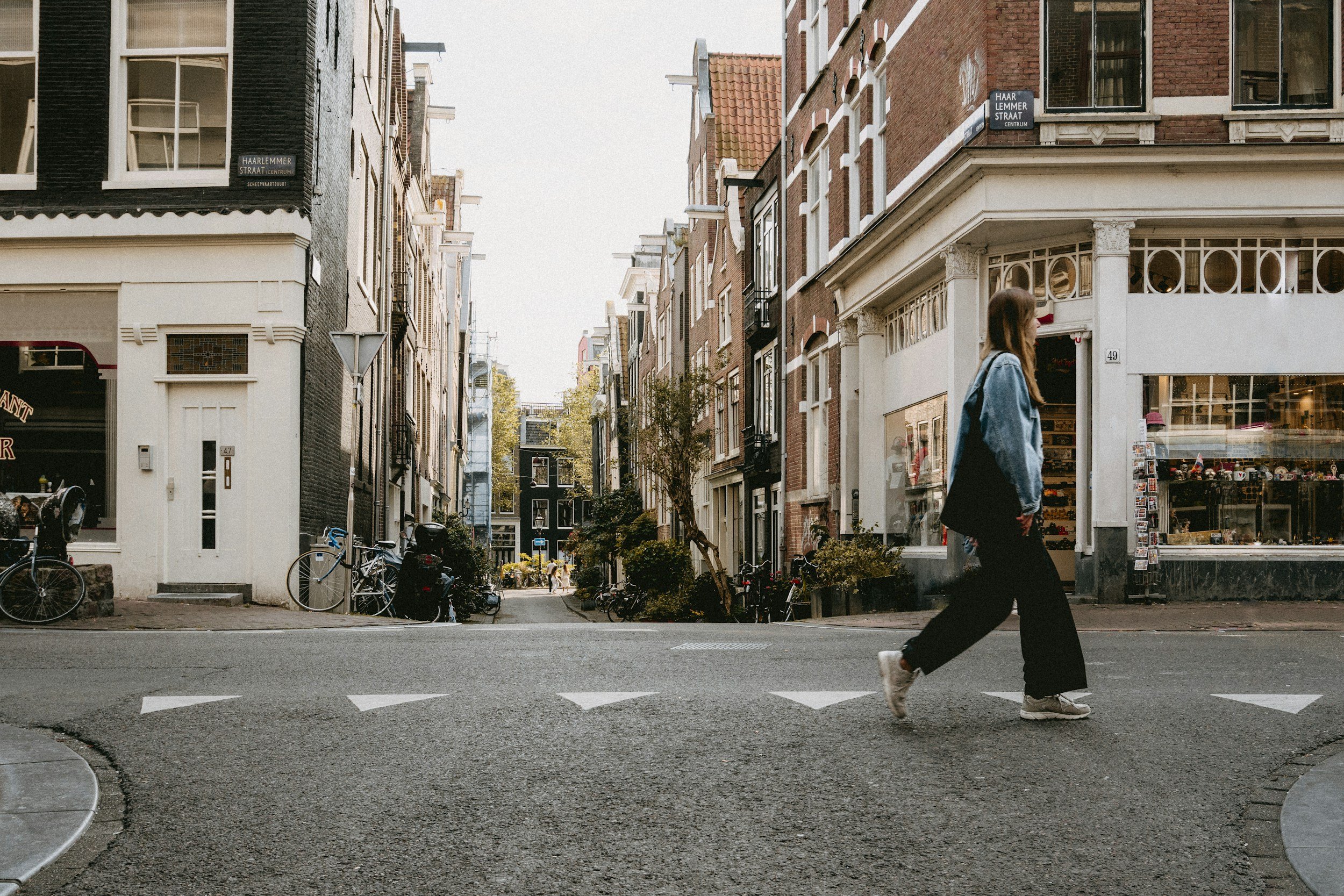 Een straat in een stad met historische gebouwen aan beide zijden. Een fiets staat geparkeerd op de stoep. Een vrouw met een denim jas en een rugzak loopt over de straat.