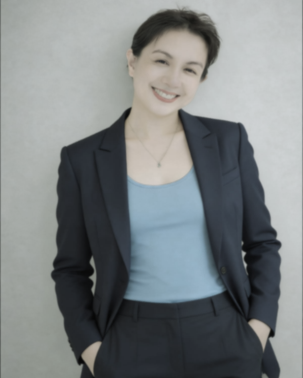 Young woman with short dark hair smiling, wearing a dark blazer over a light blue top, standing against a plain light gray background.