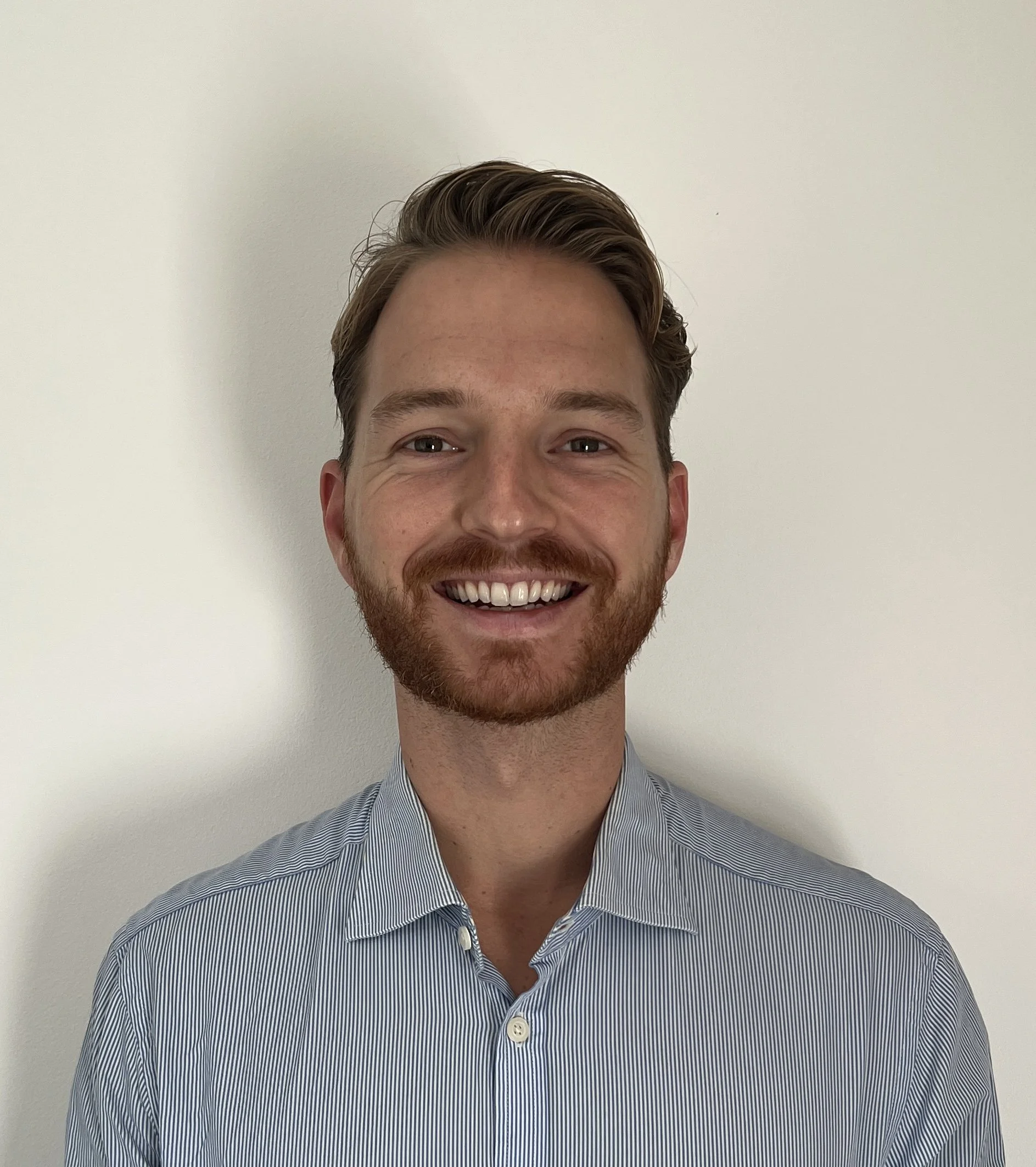 Close-up of a smiling man with brown hair and a beard wearing a blue striped button-up shirt, standing against a plain white wall.
