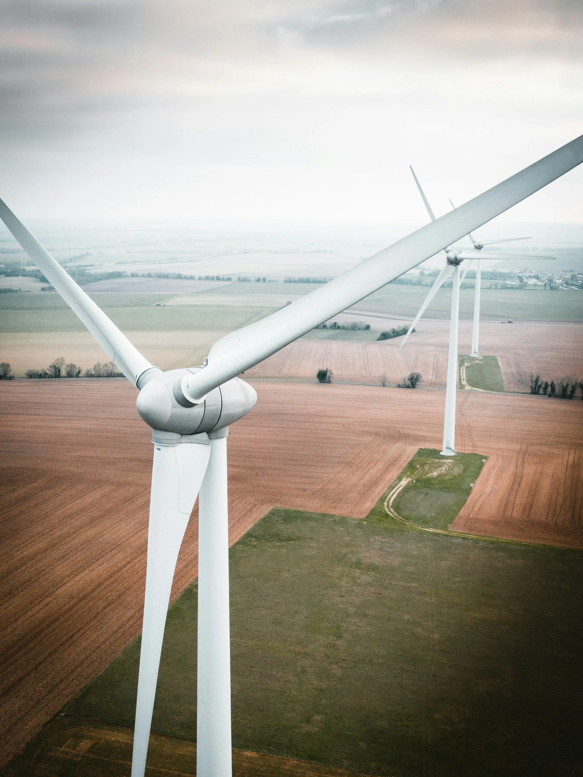 Tall white wind turbines in a rural landscape with fields and a cloudy sky.