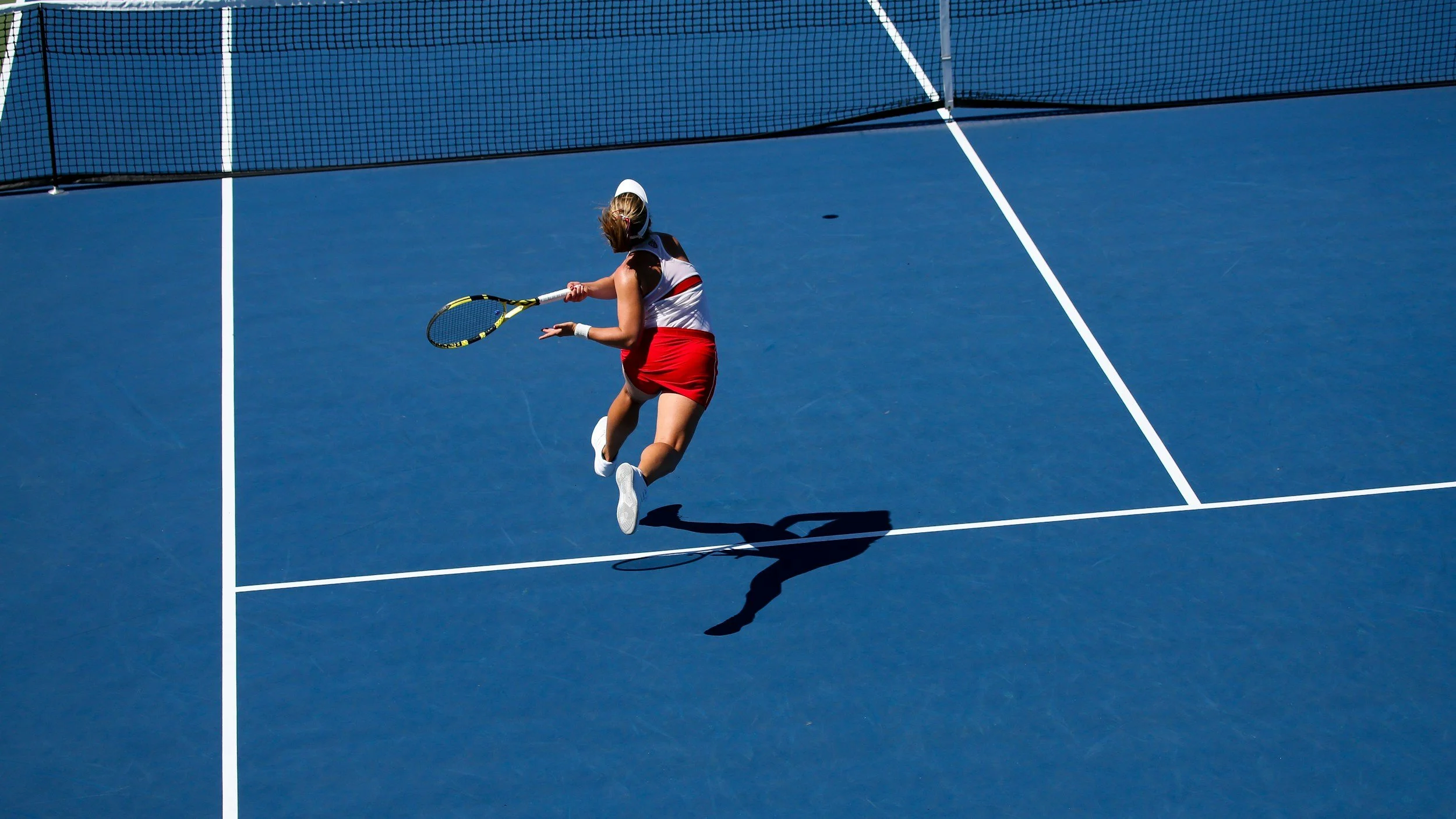 A female tennis player in a white shirt and red skirt hitting a tennis ball on a blue court with a net in the background.
