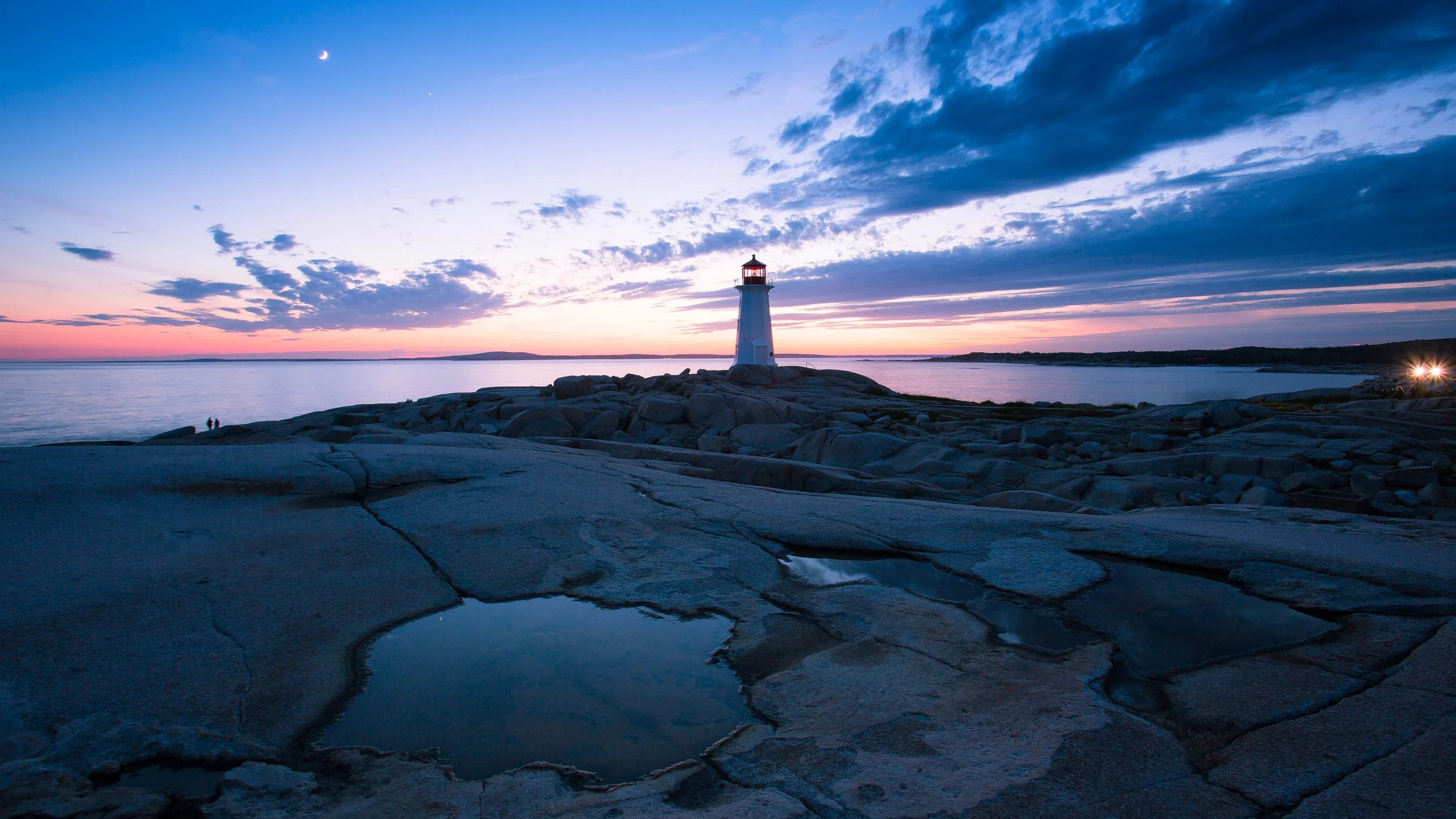 Lighthouse on rocky shoreline at sunset with colorful sky, crescent moon, and water in the background.