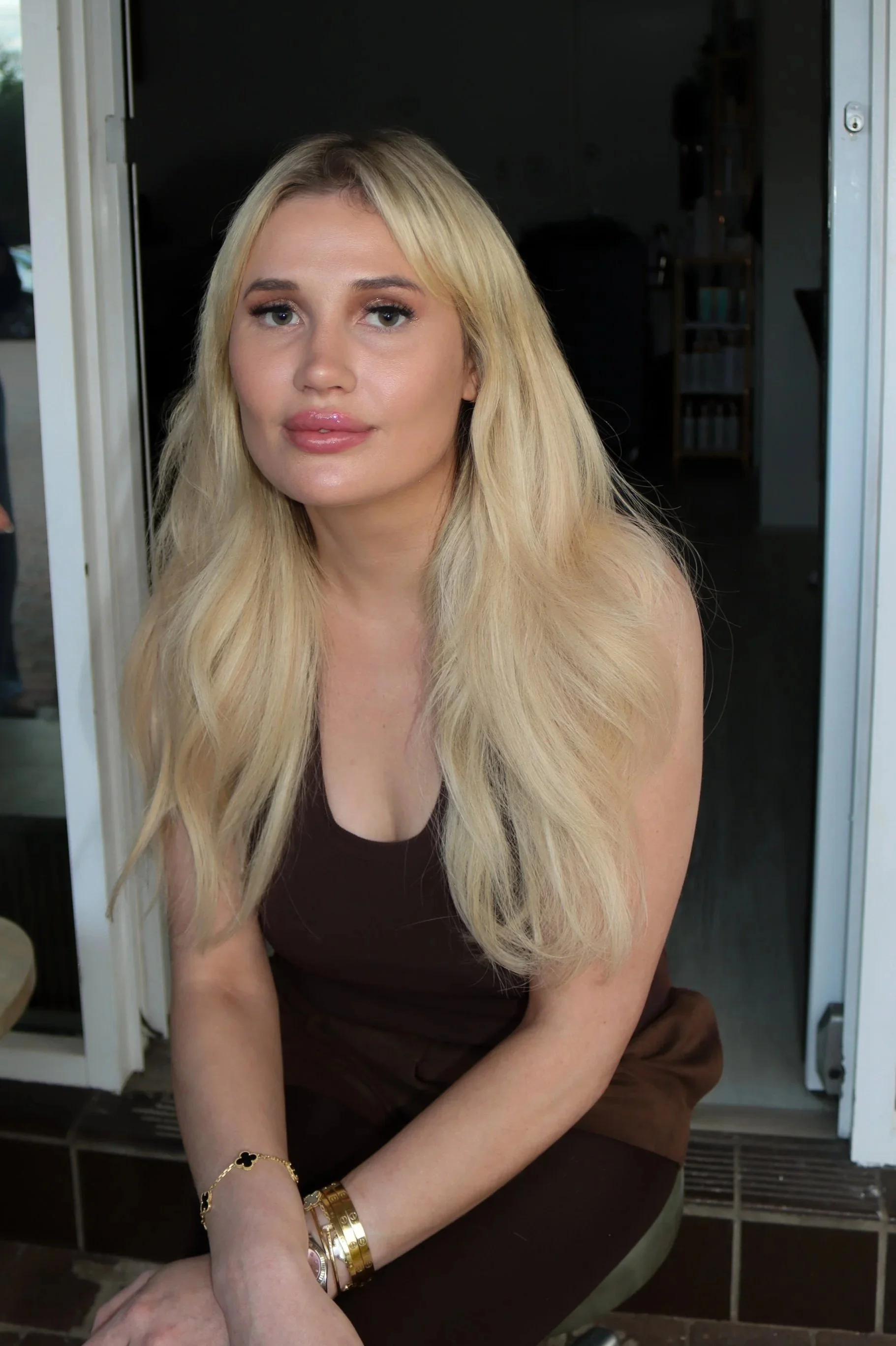 A woman with long blonde hair, wearing a dark sleeveless top, sitting on a porch with her arms crossed, wearing a gold bracelet and watch.