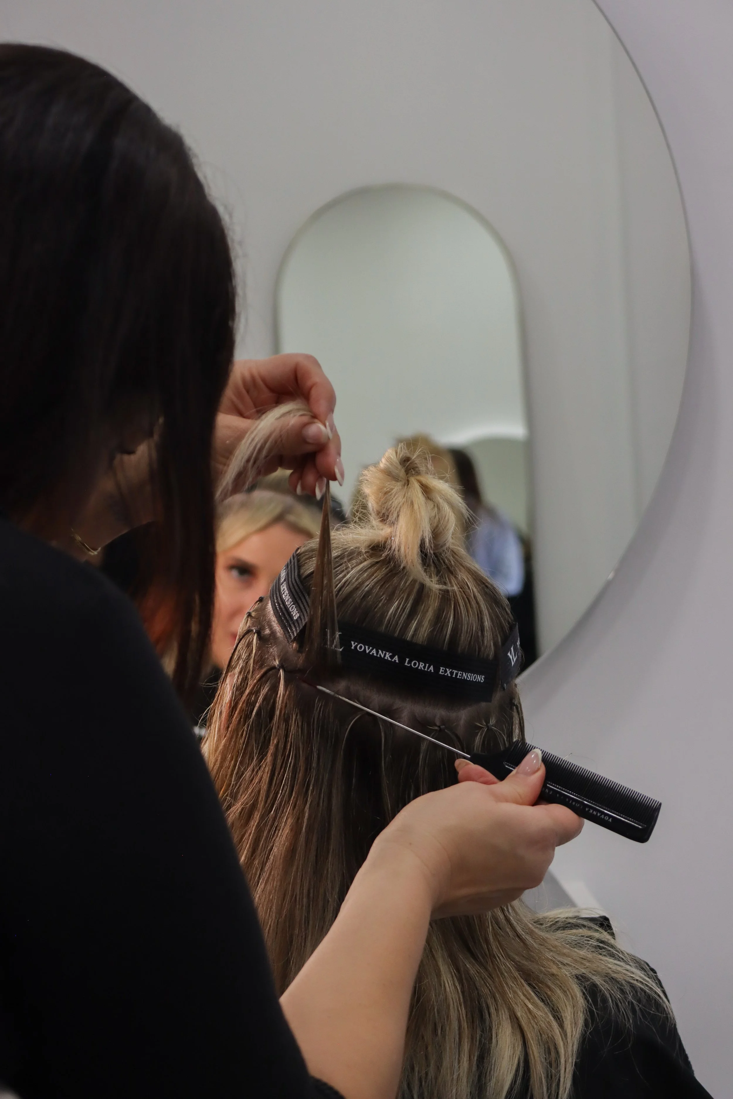 A hairstylist works on a woman's hair in front of a mirror, using a comb and scissors, while another woman looks on in a salon with white walls.