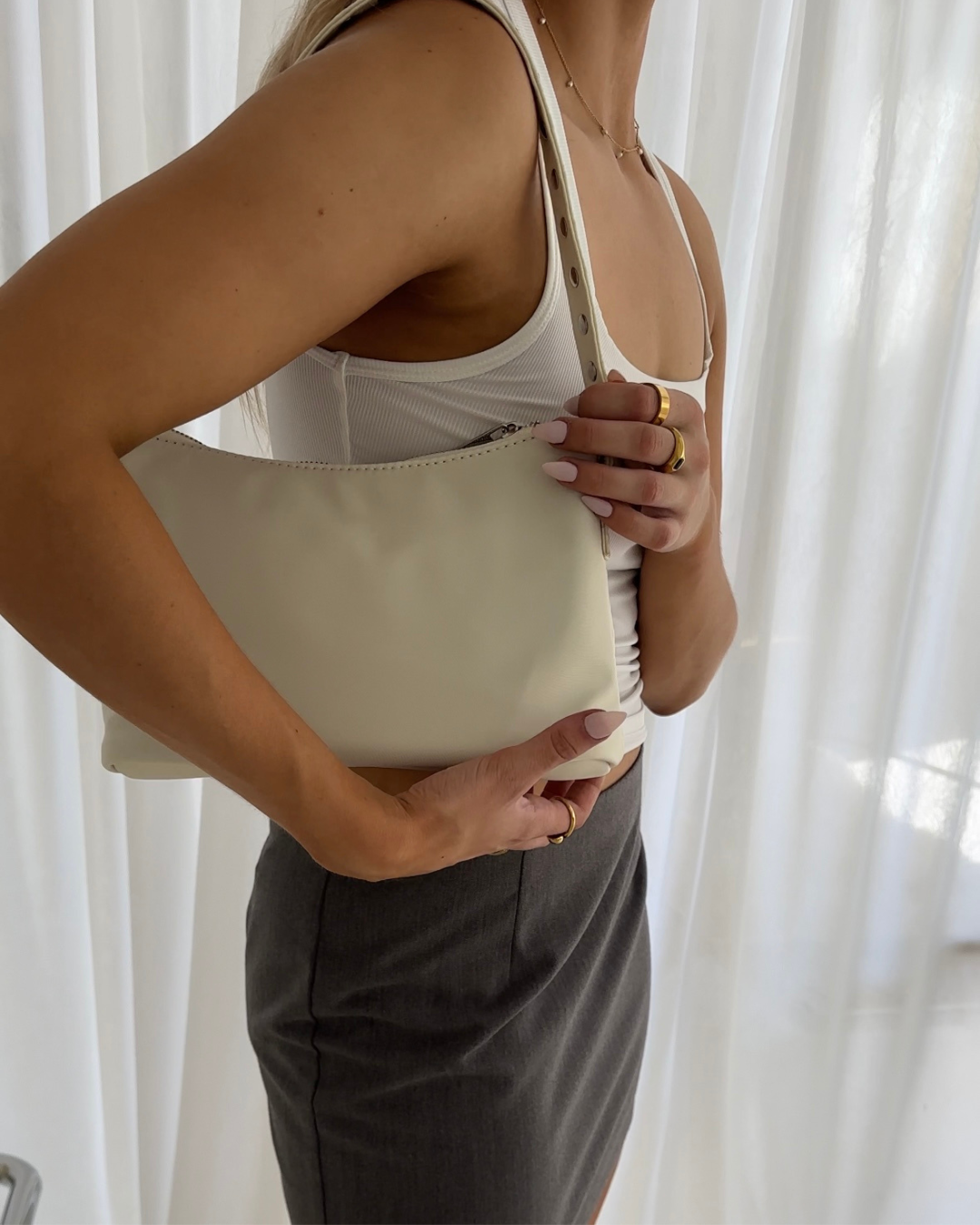 A woman in a white sleeveless top and gray pants is holding a cream-colored handbag, with her hand resting on the bag and her body turned slightly to the side in front of a white curtain background.