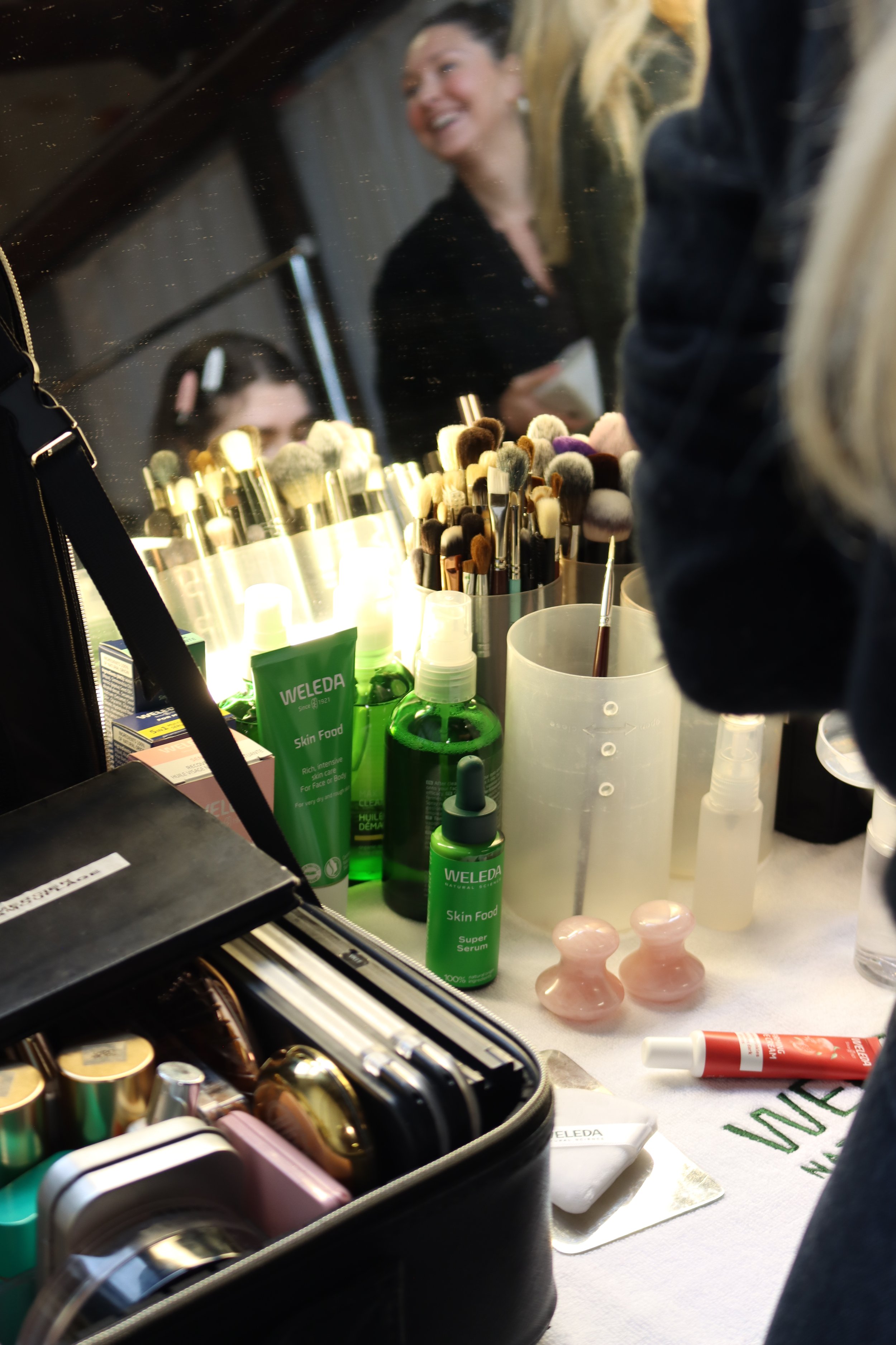 A makeup station with various cosmetics, brushes, and skincare products. A mirror reflects a smiling woman and a person with glasses. Makeup brushes are arranged in containers, and skincare items, including green bottles labeled 'Weleda,' are visible on the surface.