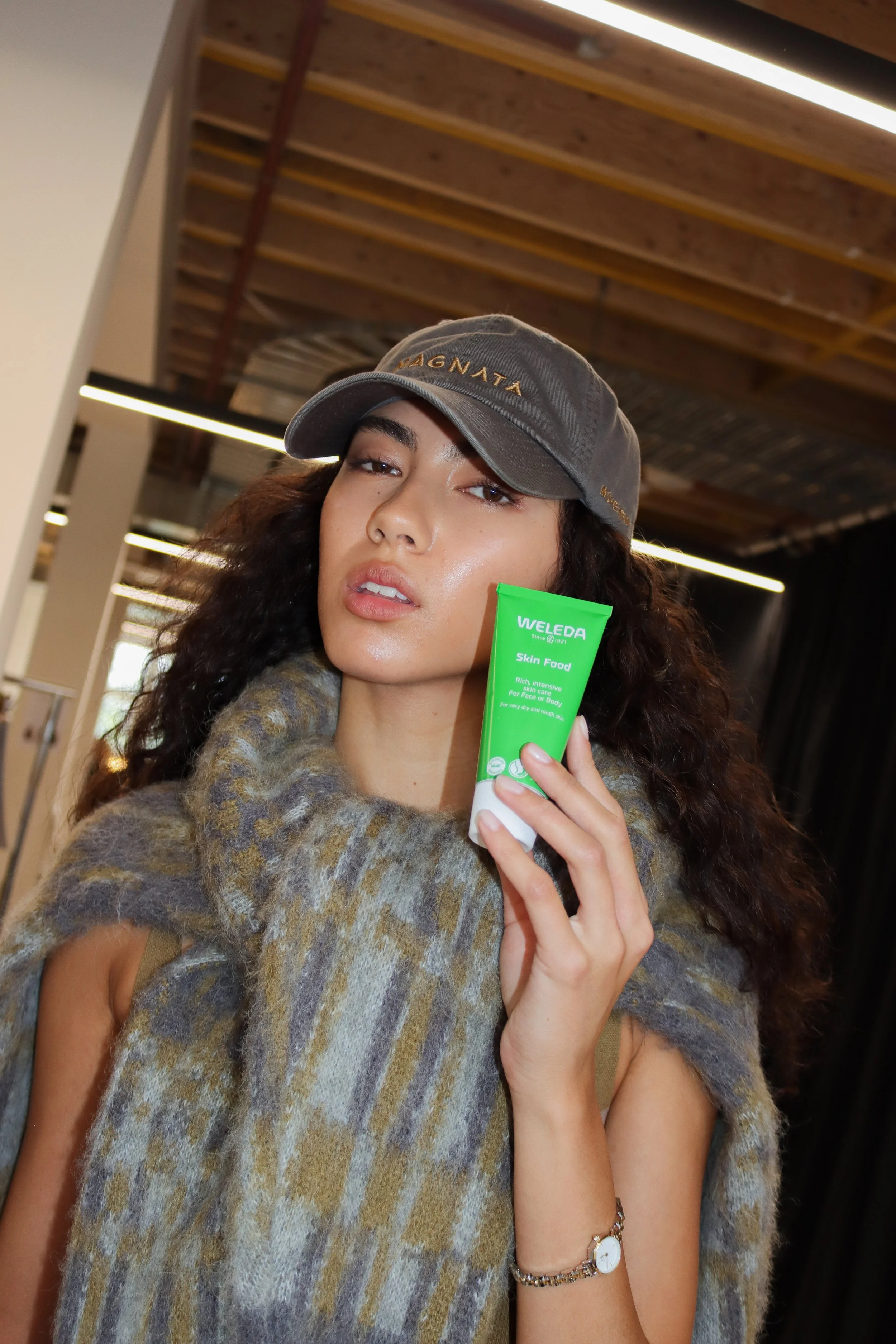 A woman with dark curly hair wearing a gray baseball cap and a multicolored fur coat, holding a green tube of Weleda Skin Food. She is indoors with a wooden ceiling and bright lighting.