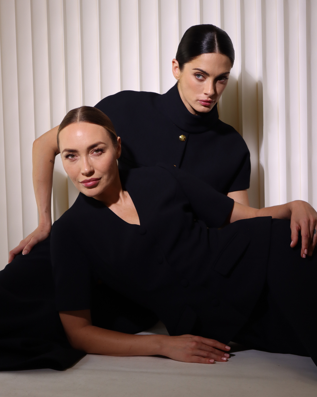 Two women with dark hair and dressed in black, posed against a white vertical blinds background.