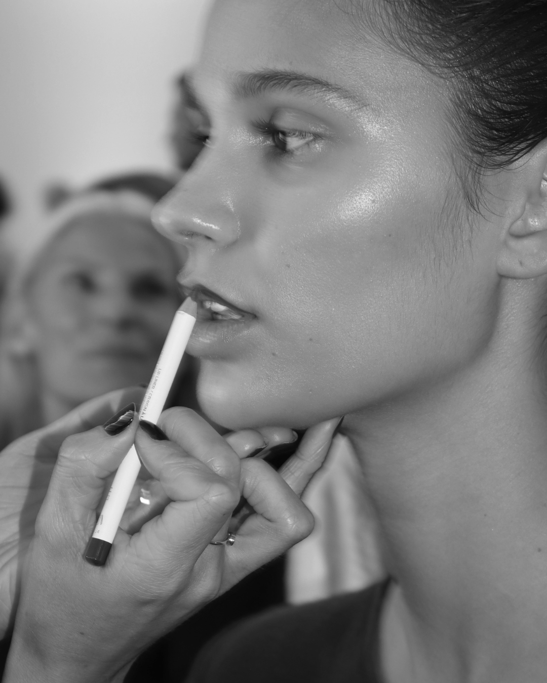 A woman getting her makeup done, with a makeup artist applying lipstick and a person in the background observing.