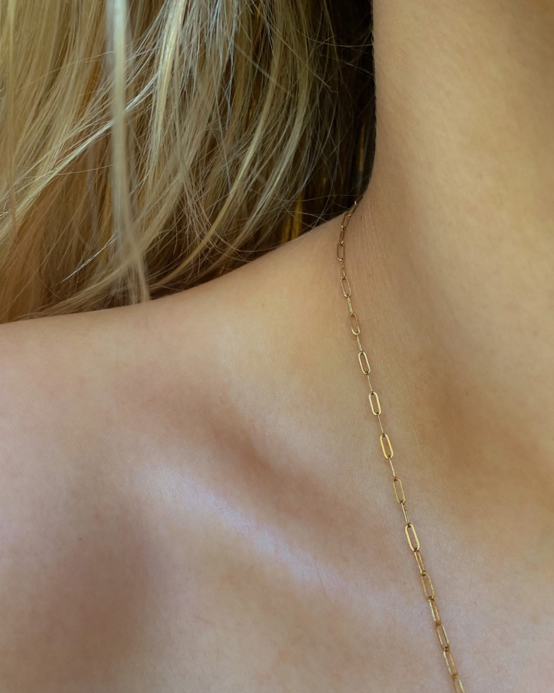 Close-up of a woman's shoulder and neck wearing a delicate gold chain necklace, with blonde hair partially covering the neck area.