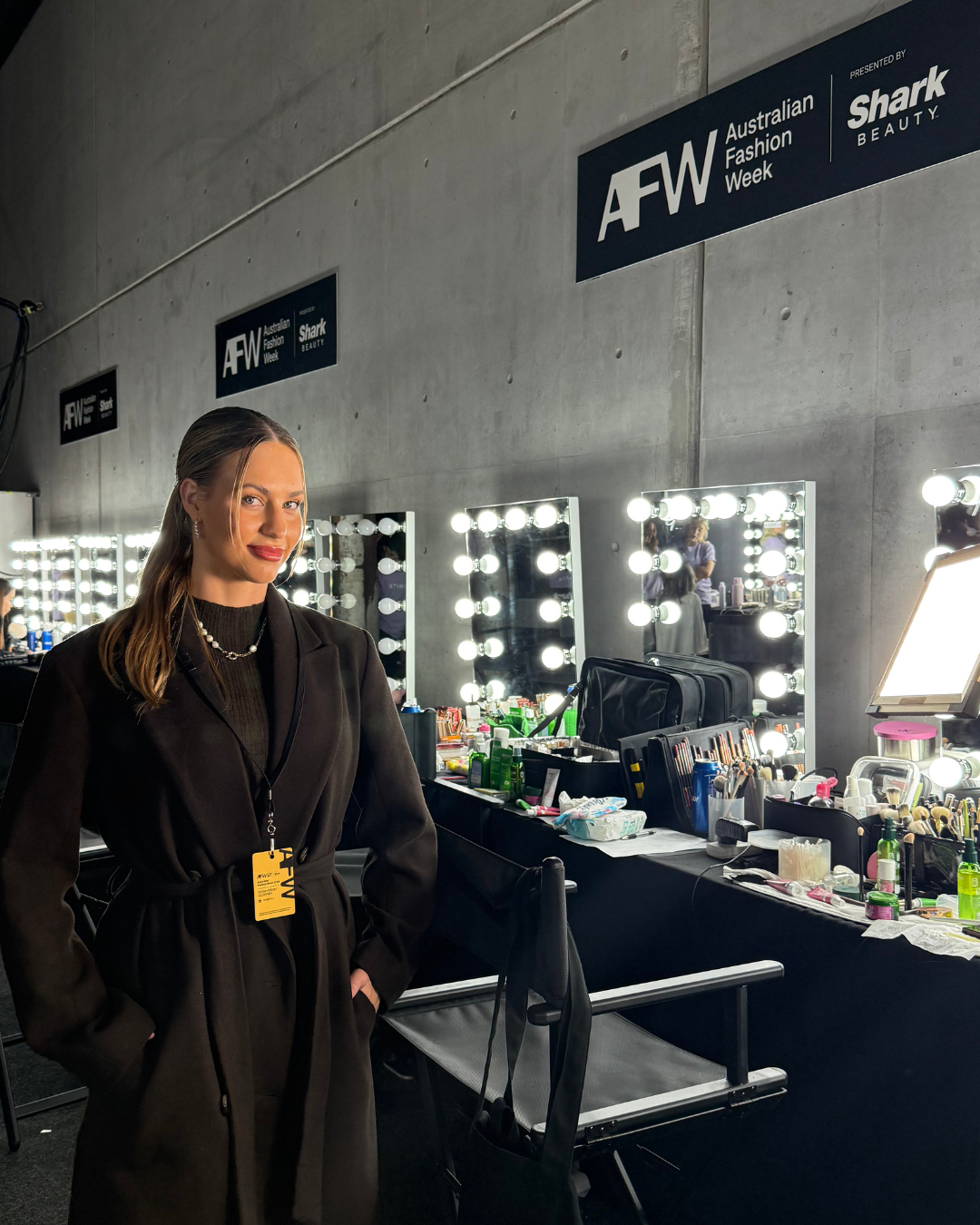 A woman in a black coat standing in front of a makeup mirror with bright lights backstage at Australian Fashion Week.