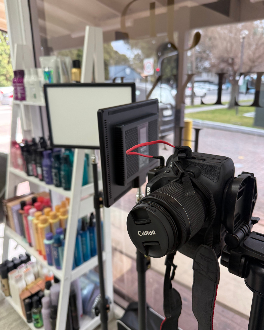 A Canon camera mounted on a tripod with a mounted monitor, set up to film or photograph inside a store, with shelves of products in the background and a storefront window showing outside scenery.