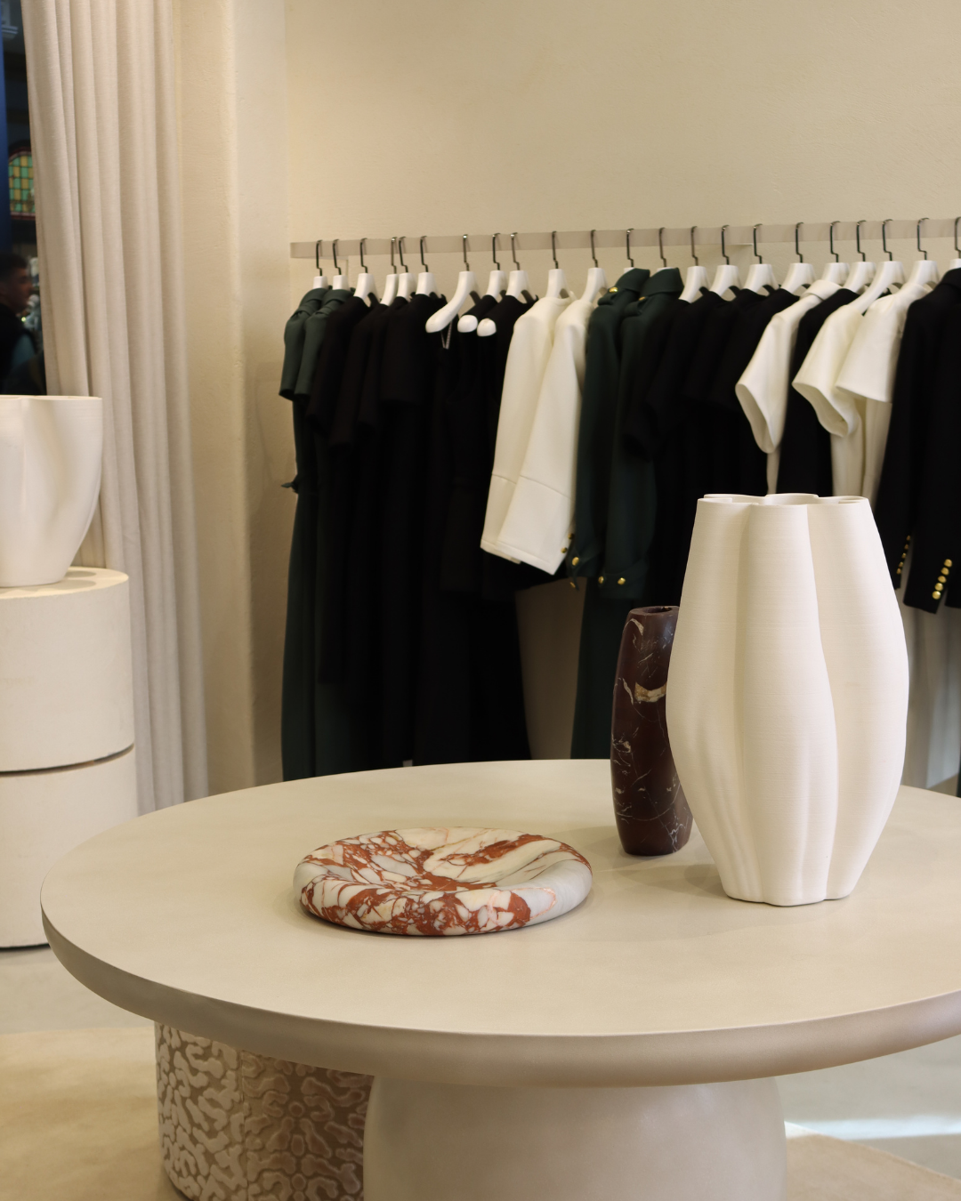 Clothing rack with black, white, and dark green shirts and jackets, along with decorative vases and a marble dish on a round table in a store or showroom.