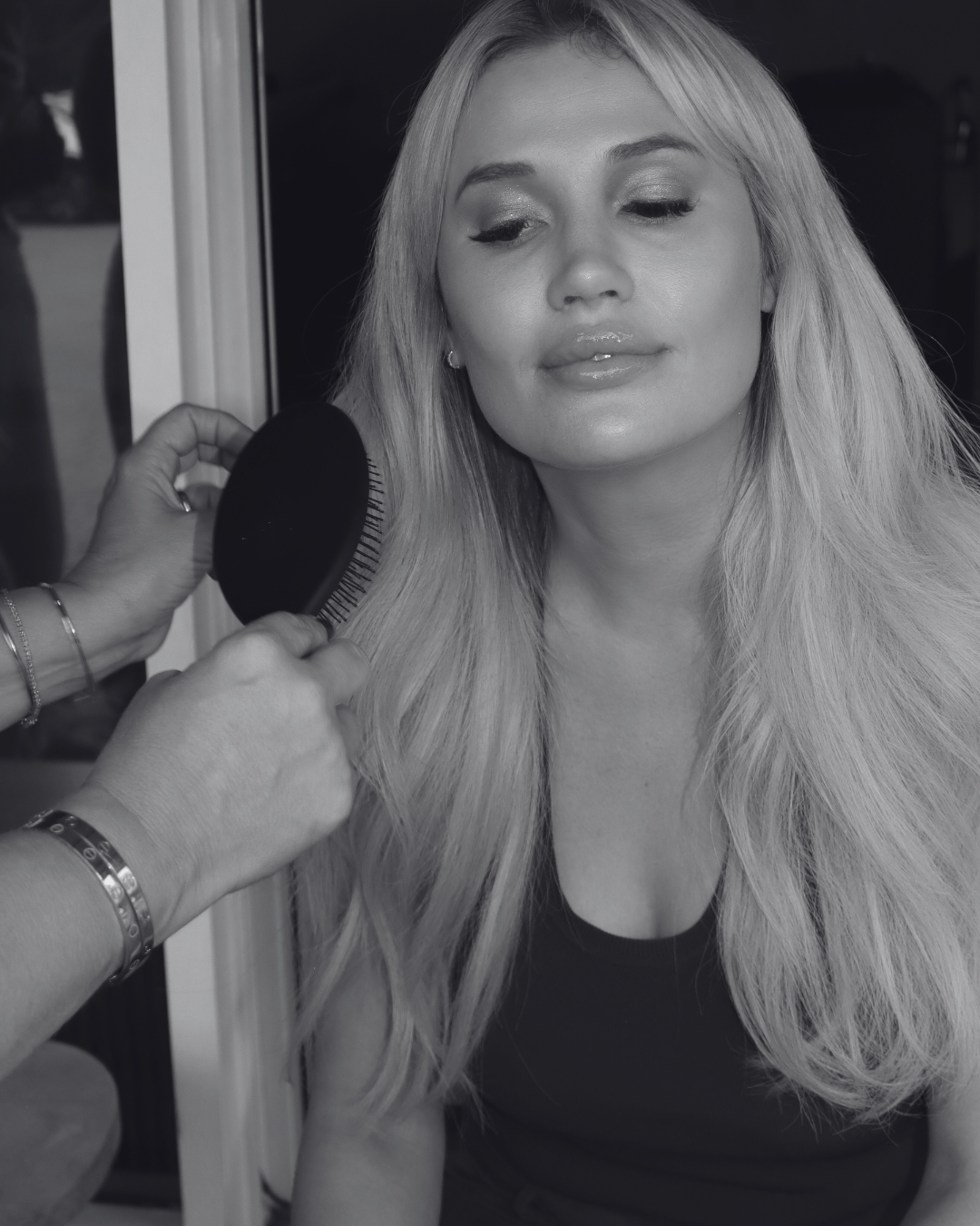 A woman with long blonde hair sitting as a person brushes her hair with a round hairbrush.