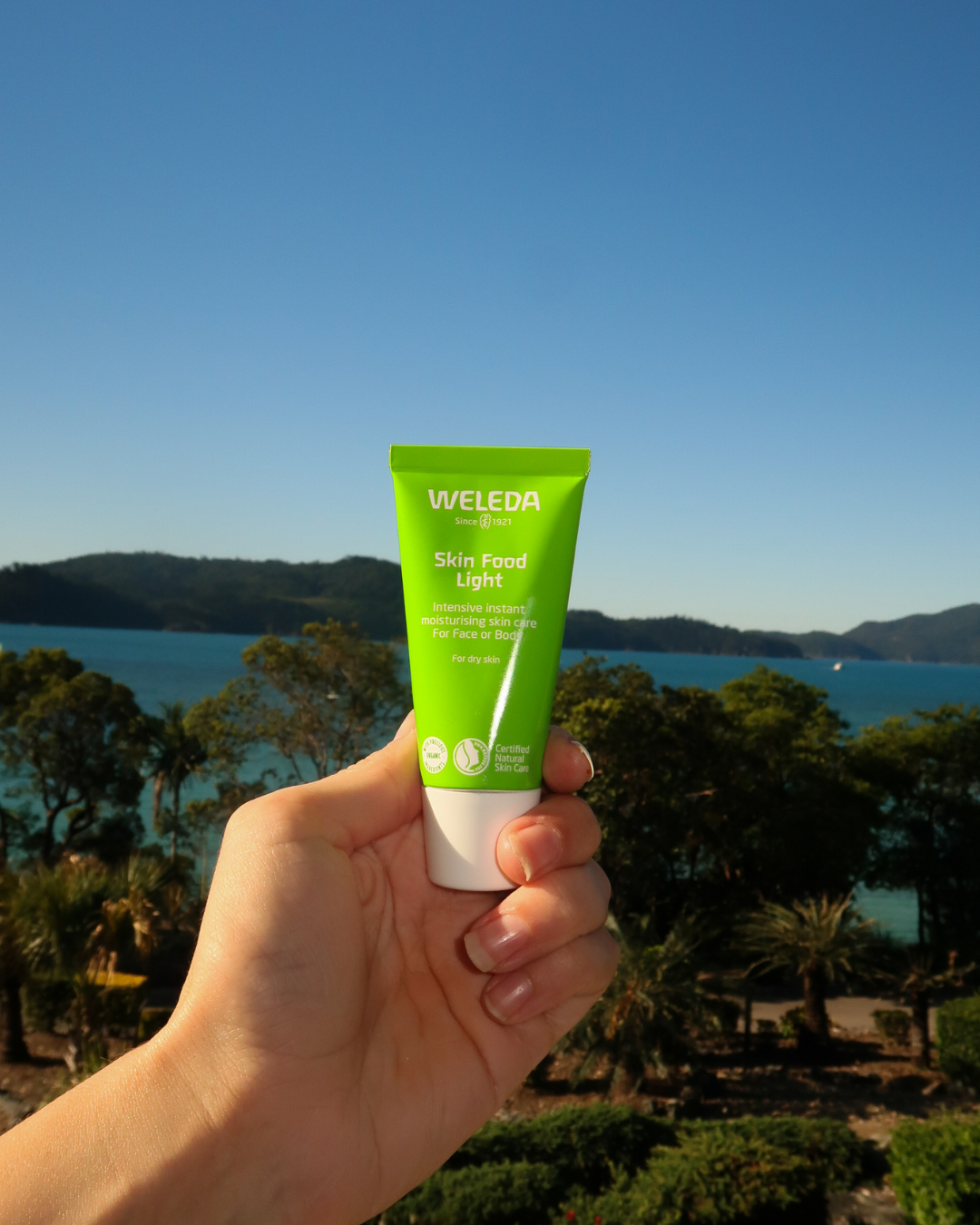 Hand holding a green tube of Weleda Skin Food Light moisturizer against a scenic background of trees, water, and mountains under a clear blue sky.