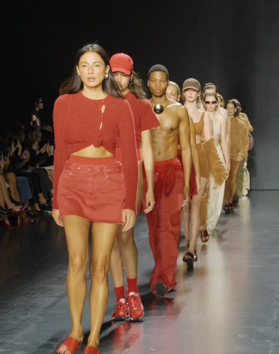 Models walking down a runway during a fashion show, wearing red and neutral-colored outfits.