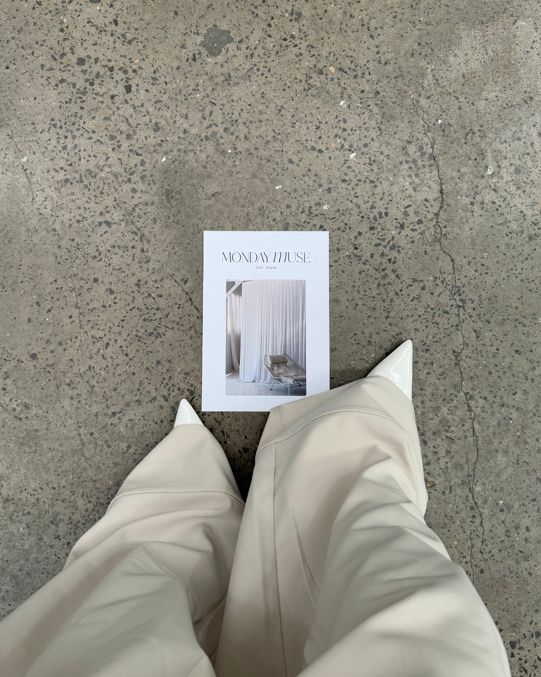 Photo from a high perspective showing the person's white pointed-toe shoes, beige pants, and a magazine or brochure titled 'Monday Muse' on a concrete floor with a cracked line.