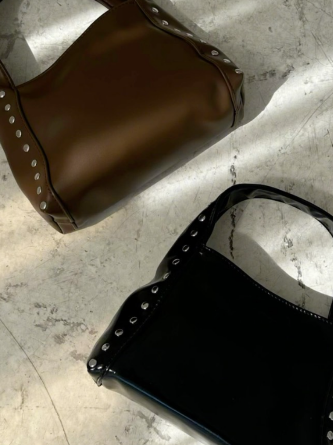 Close-up of two glossy handbags, one brown and one black, on a concrete floor, each featuring silver studs along the edges.