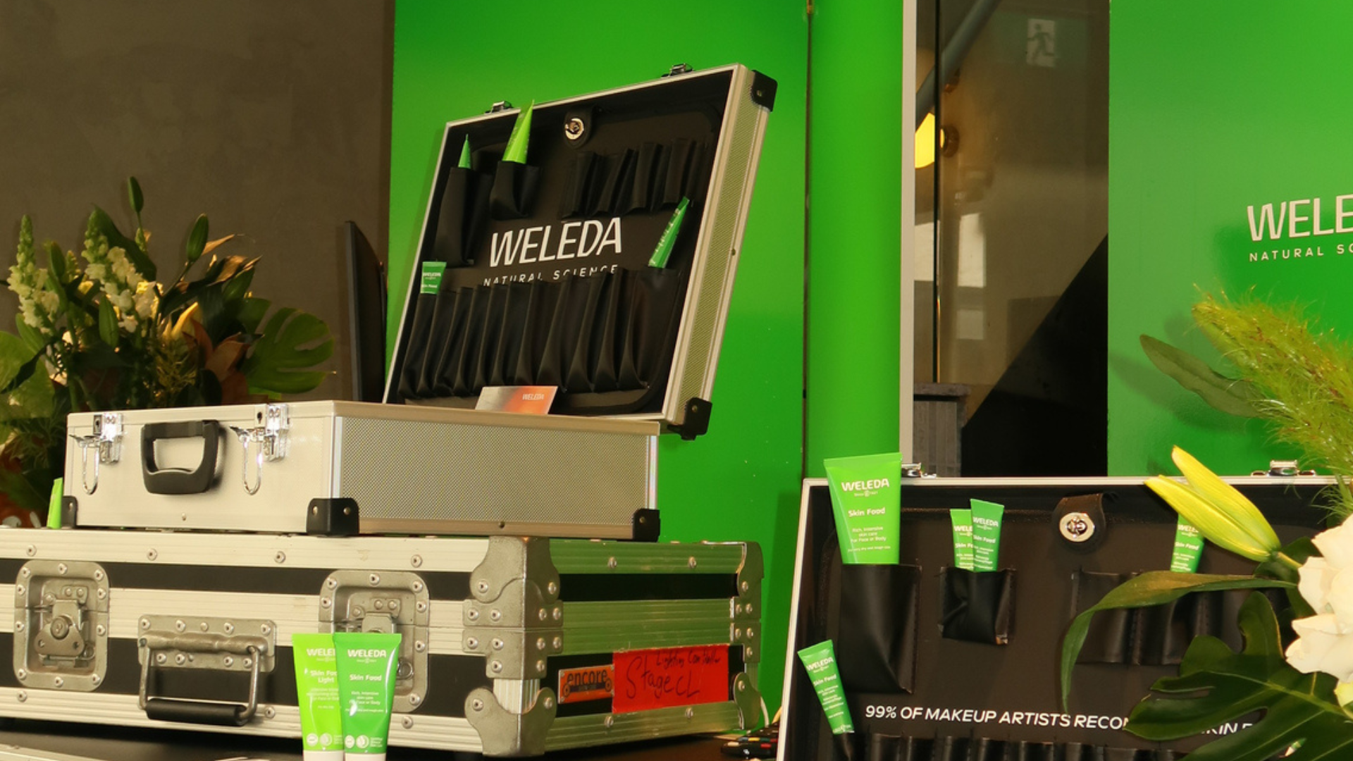 A display of Weleda natural skincare products arranged on a table with some opened cases, green background, and flowers on the side.