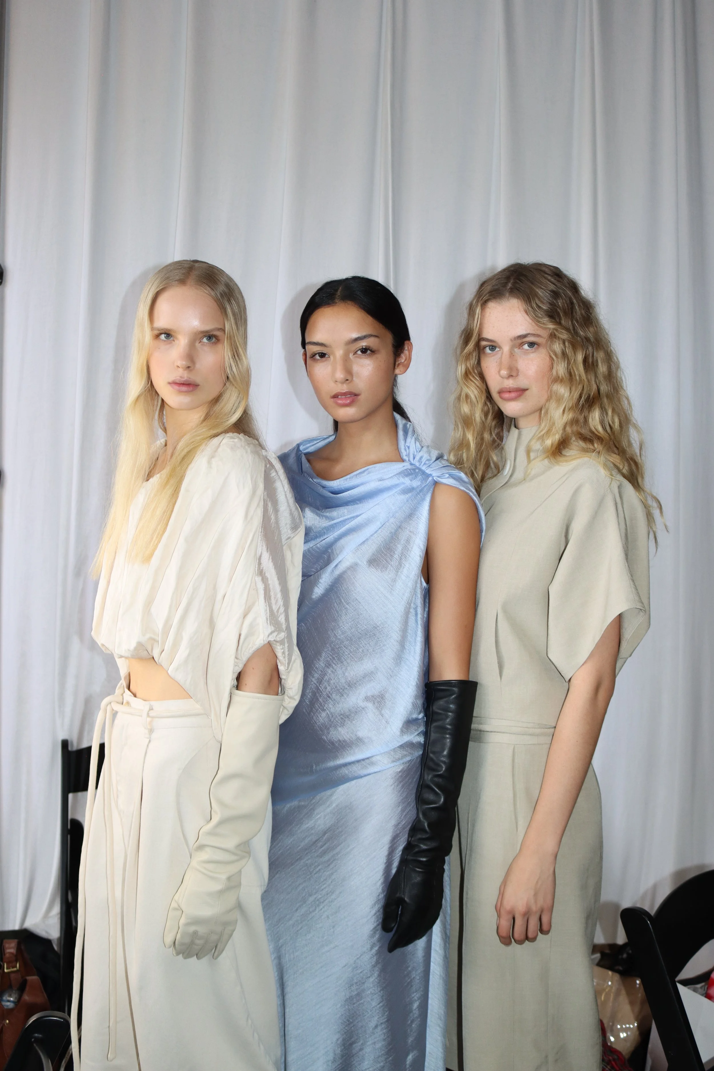Three women standing together against a white curtain backdrop, dressed in cream, light blue, and beige outfits, with some wearing gloves and arm accessories, posing confidently.