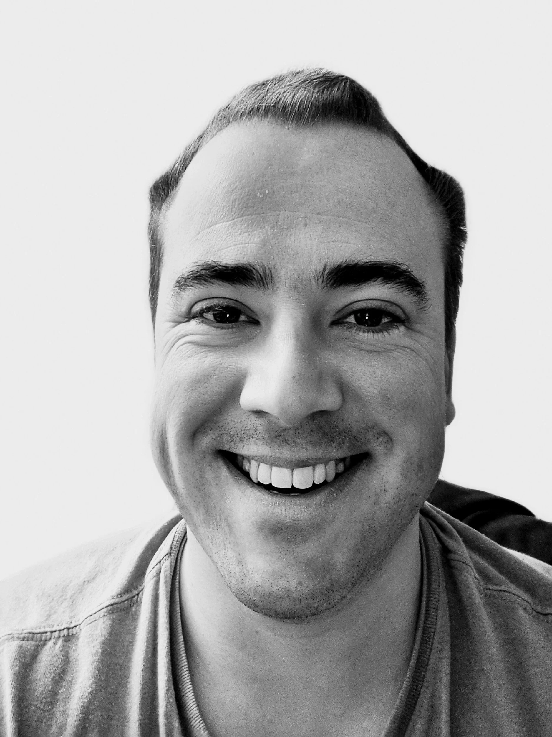 A smiling man with short dark hair, wearing a t-shirt, taking a selfie in black and white.