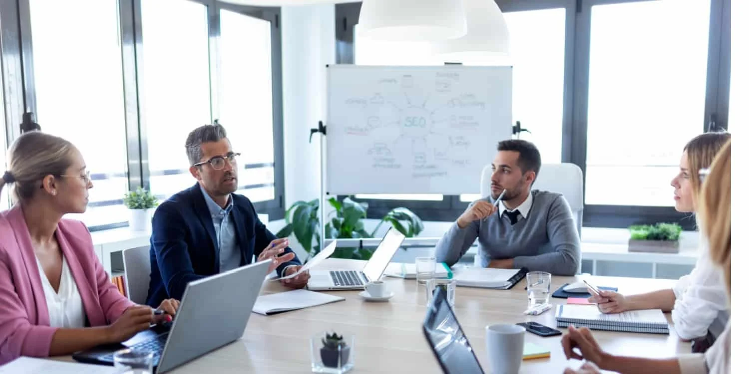 People sitting around a conference table in an office, engaged in a discussion. There are laptops, notebooks, and a whiteboard with notes behind them.