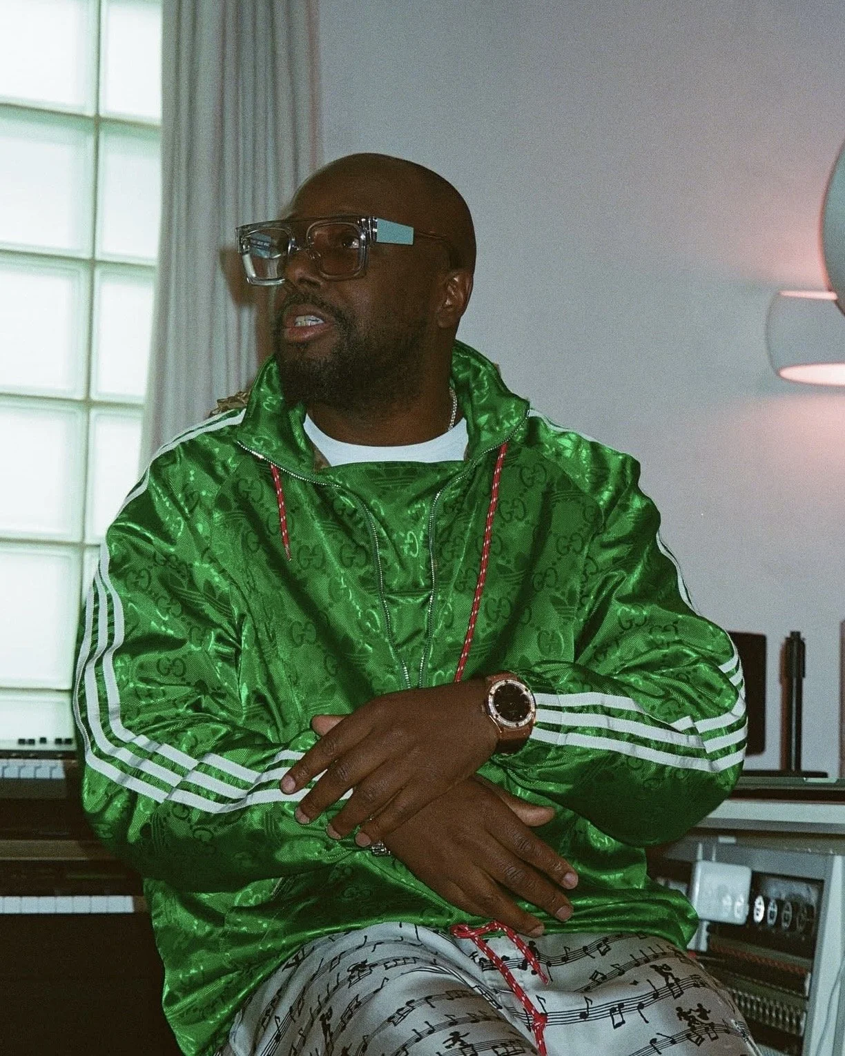 A man wearing a green Gucci jacket, white music note pants, and glasses sitting in front of a digital piano in a room with natural light from large windows.