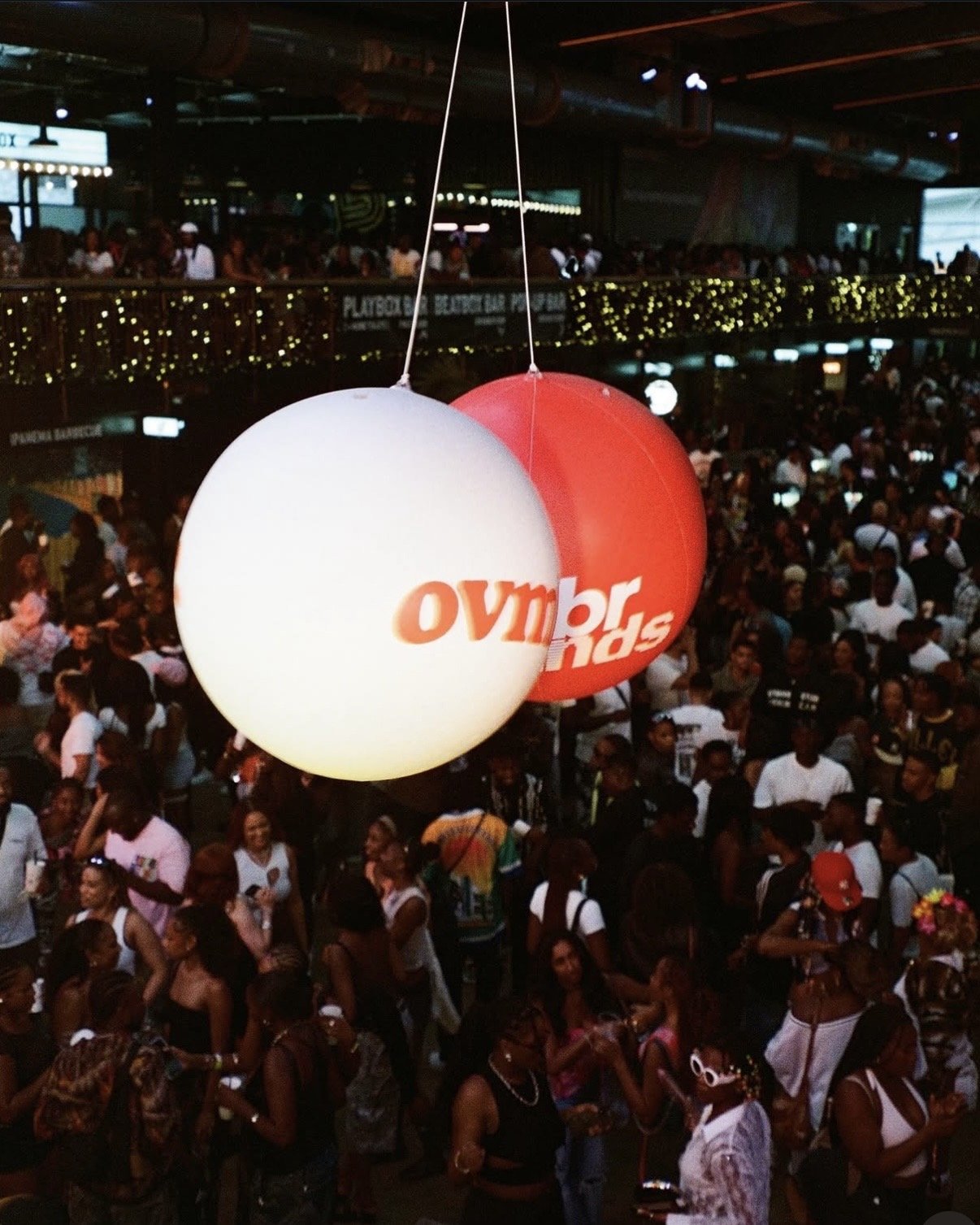 A crowded indoor event with two hanging balloons, one white with orange text and one red with white text, surrounded by numerous people.