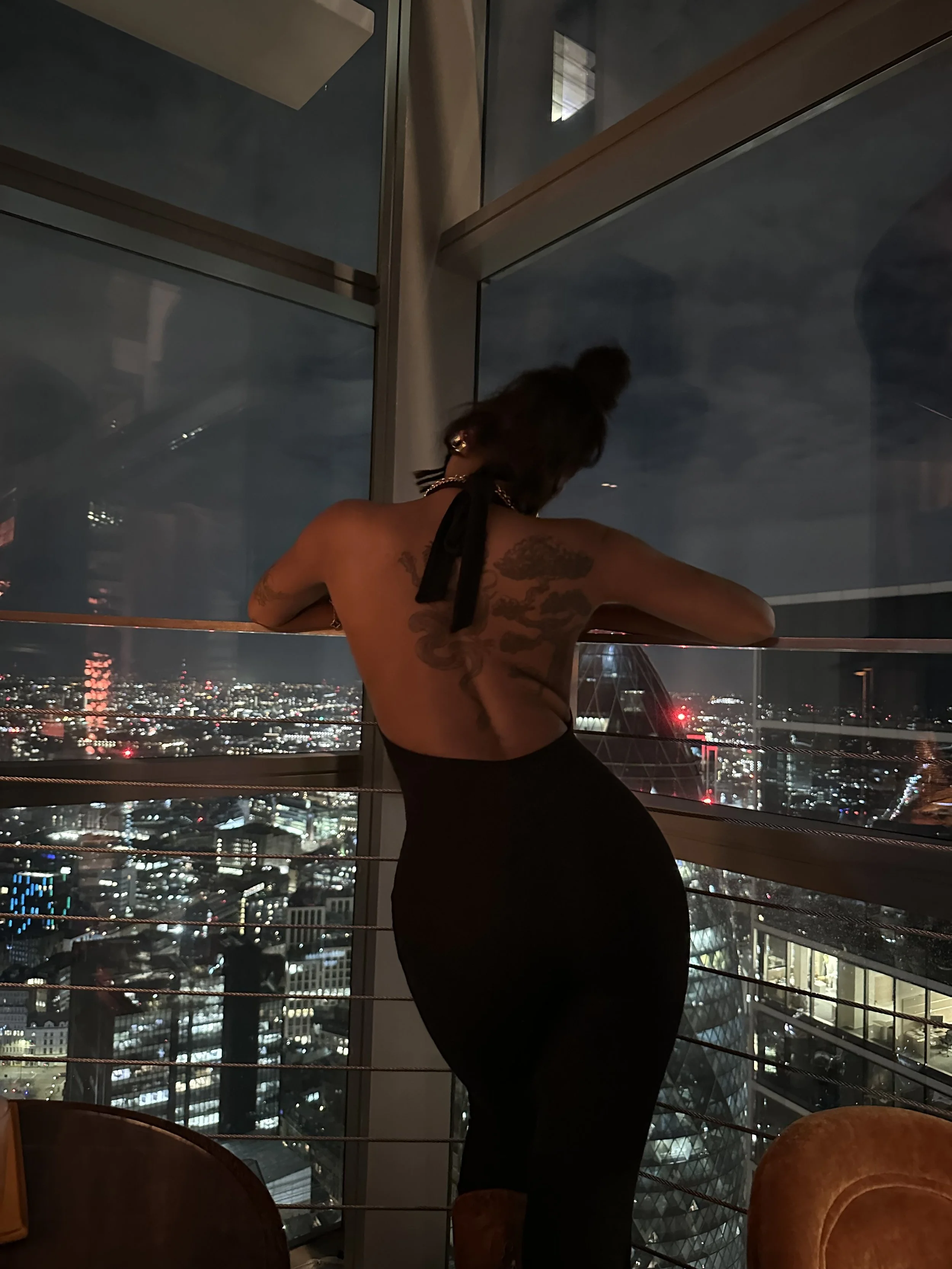 A woman with tattoos and a bun hairstyle leaning on a railing inside a tall building, looking at a city skyline at night with lights and skyscrapers visible outside the glass windows.