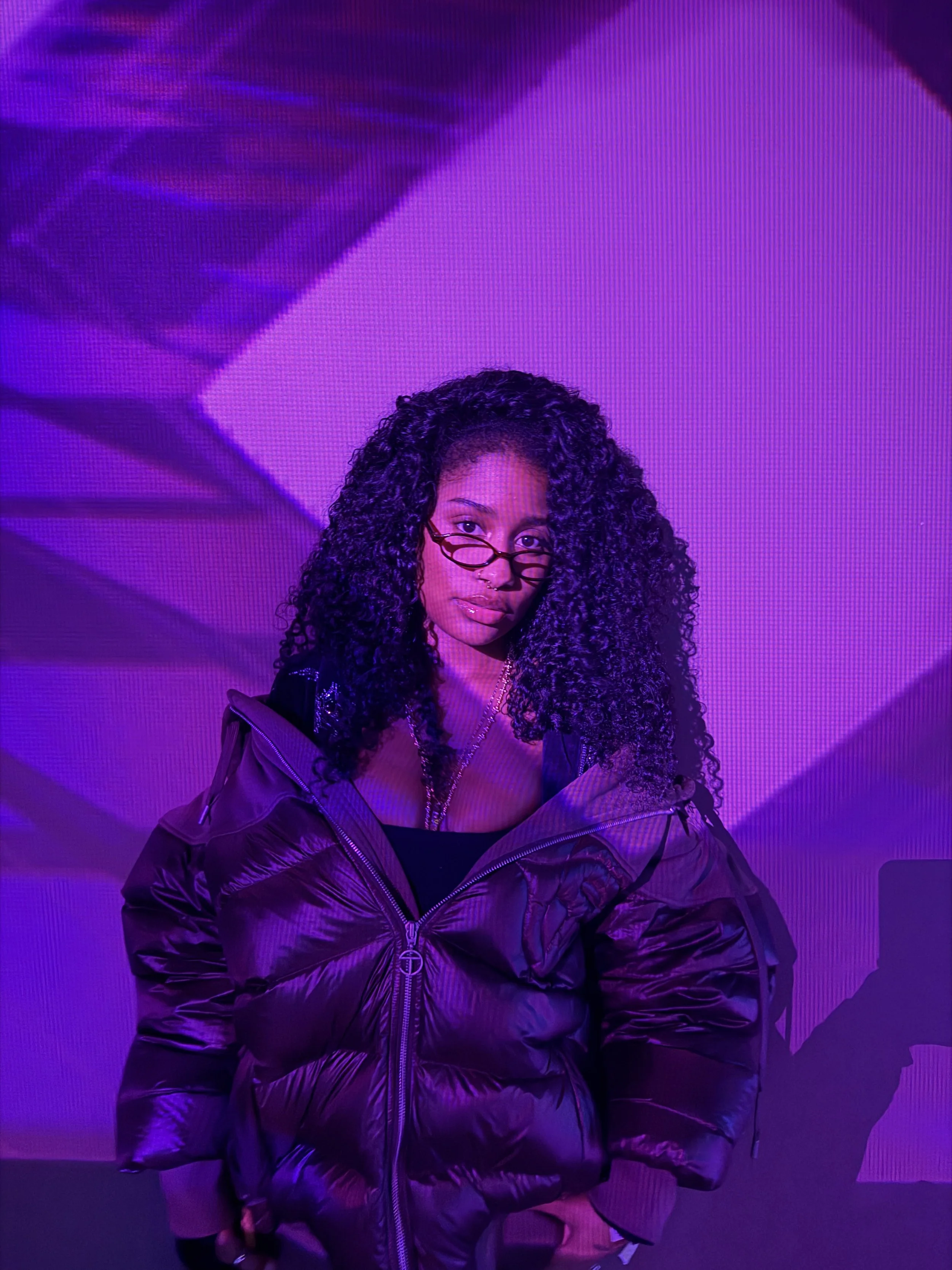 A woman with curly hair, wearing glasses and a dark puffer jacket, stands in front of a purple and pink LED-lit background.