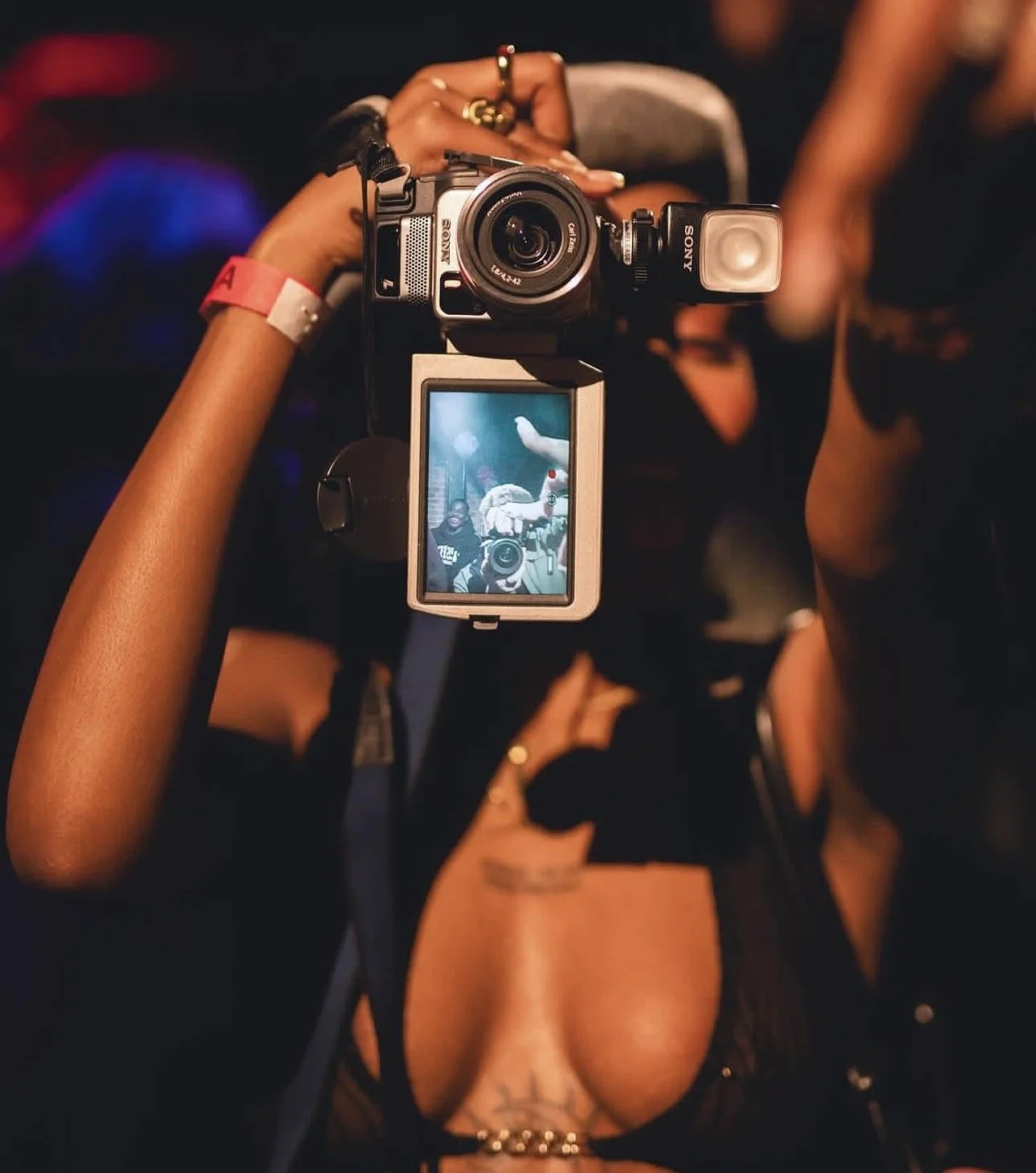 Person holding a camera with a flip-out screen showing their reflection, in a dimly lit setting with blurred background.