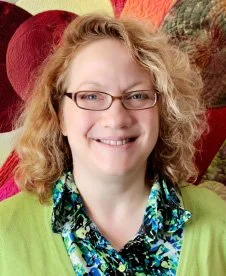 Smiling woman with curly blonde hair and glasses, wearing a colorful patterned shirt and green cardigan, in front of a red and pink background.