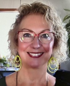 Close-up of a woman with curly blonde hair, wearing glasses and yellow hoop earrings, smiling indoors with plants in the background.