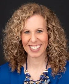 Portrait of a woman with curly blonde hair wearing a blue blazer and a matching necklace, smiling against a dark background.