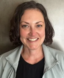Close-up of a smiling woman with short, wavy dark hair wearing a gray jacket and black top.