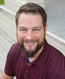A smiling man with a beard and short dark hair, wearing a maroon polo shirt, sitting outside on steps.